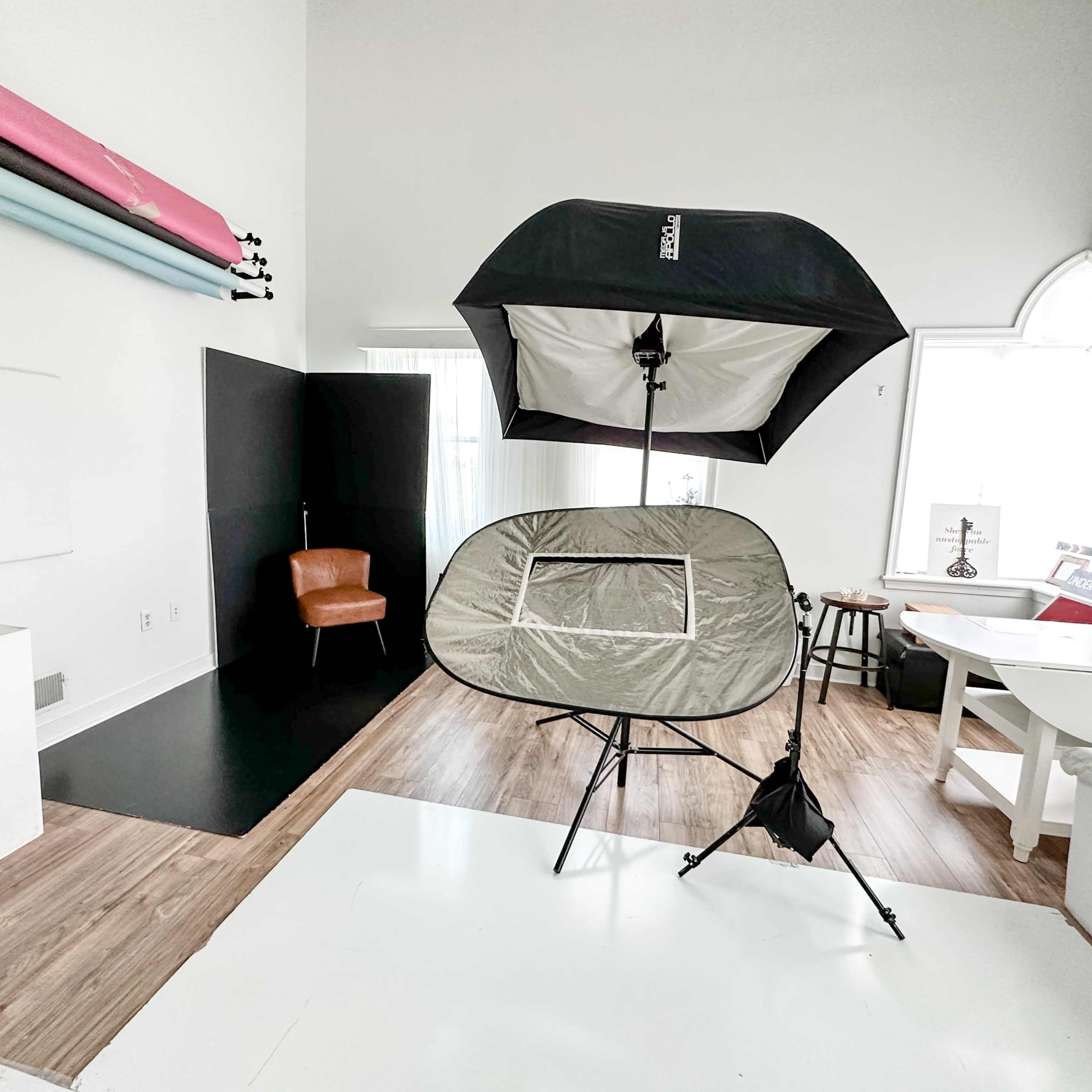 A photography studio setup featuring a light umbrella, a portable backdrop, a wooden chair, and various backdrops on the wall.