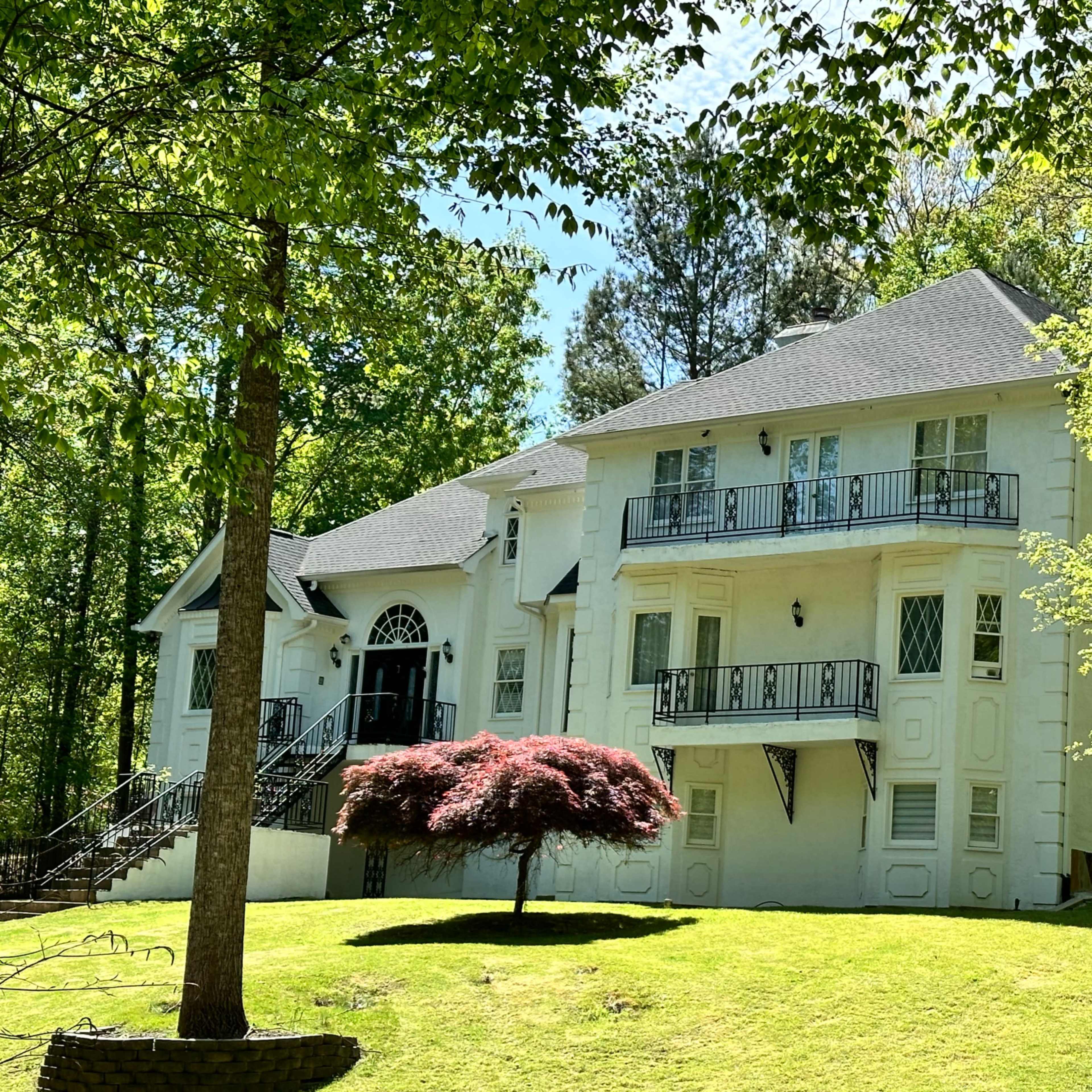 A two-story white house with a symmetrical facade and balconies is set against a backdrop of green trees and a manicured lawn.