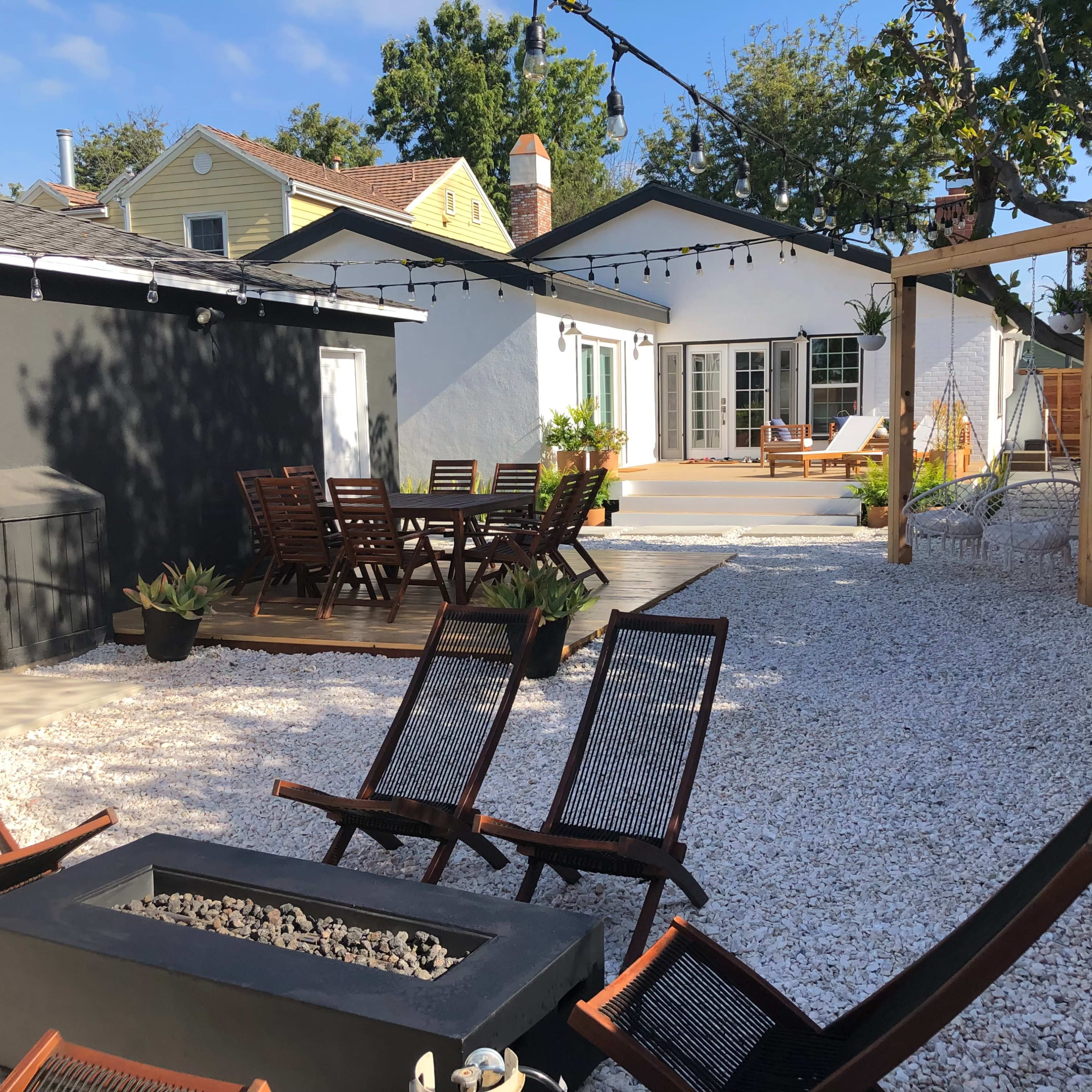 A backyard with a gravel surface, a seating area with wooden furniture, a fire pit, and a house with a patio in the background.