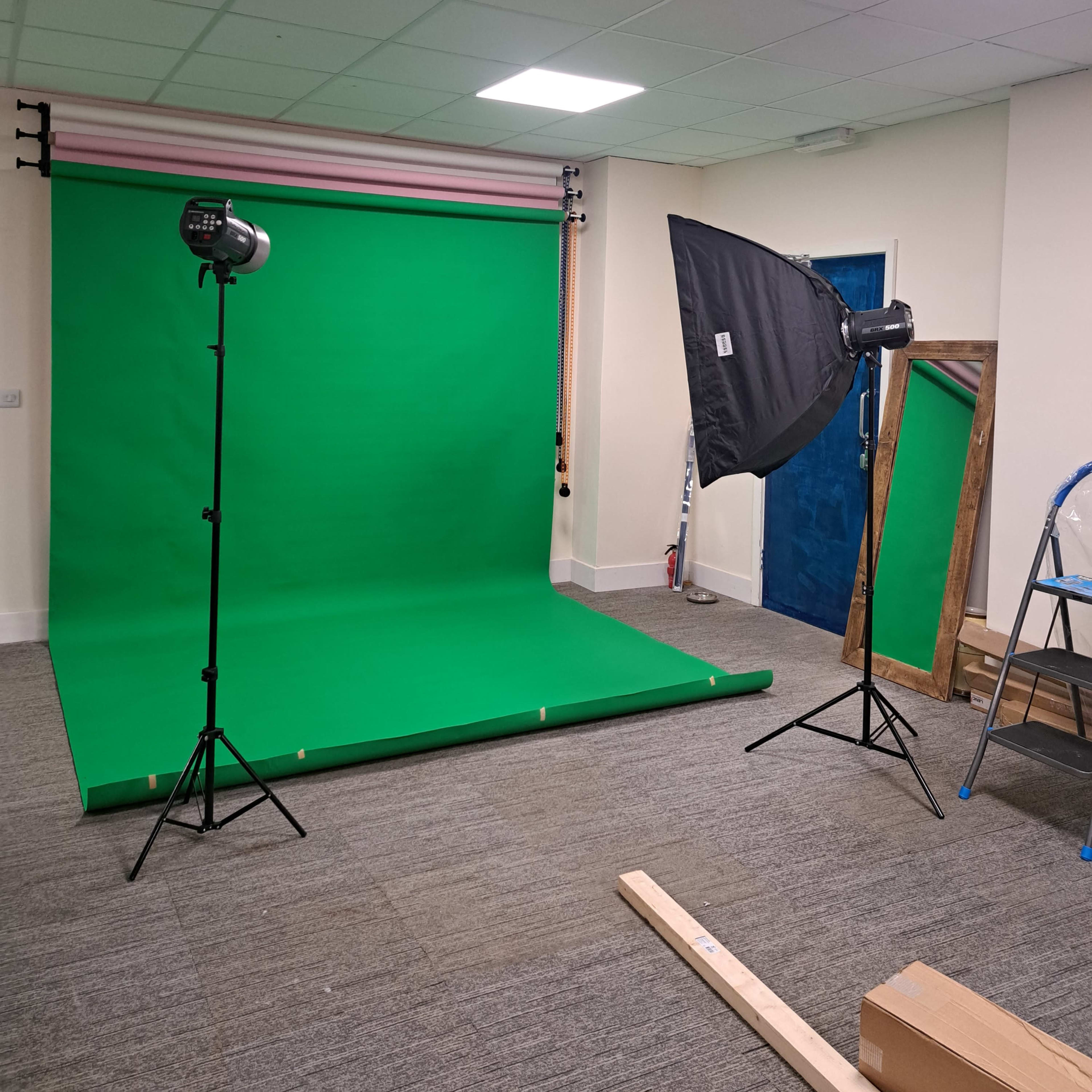 A photography studio setup with a green backdrop, two light stands, and a large mirror in a room with gray carpet.