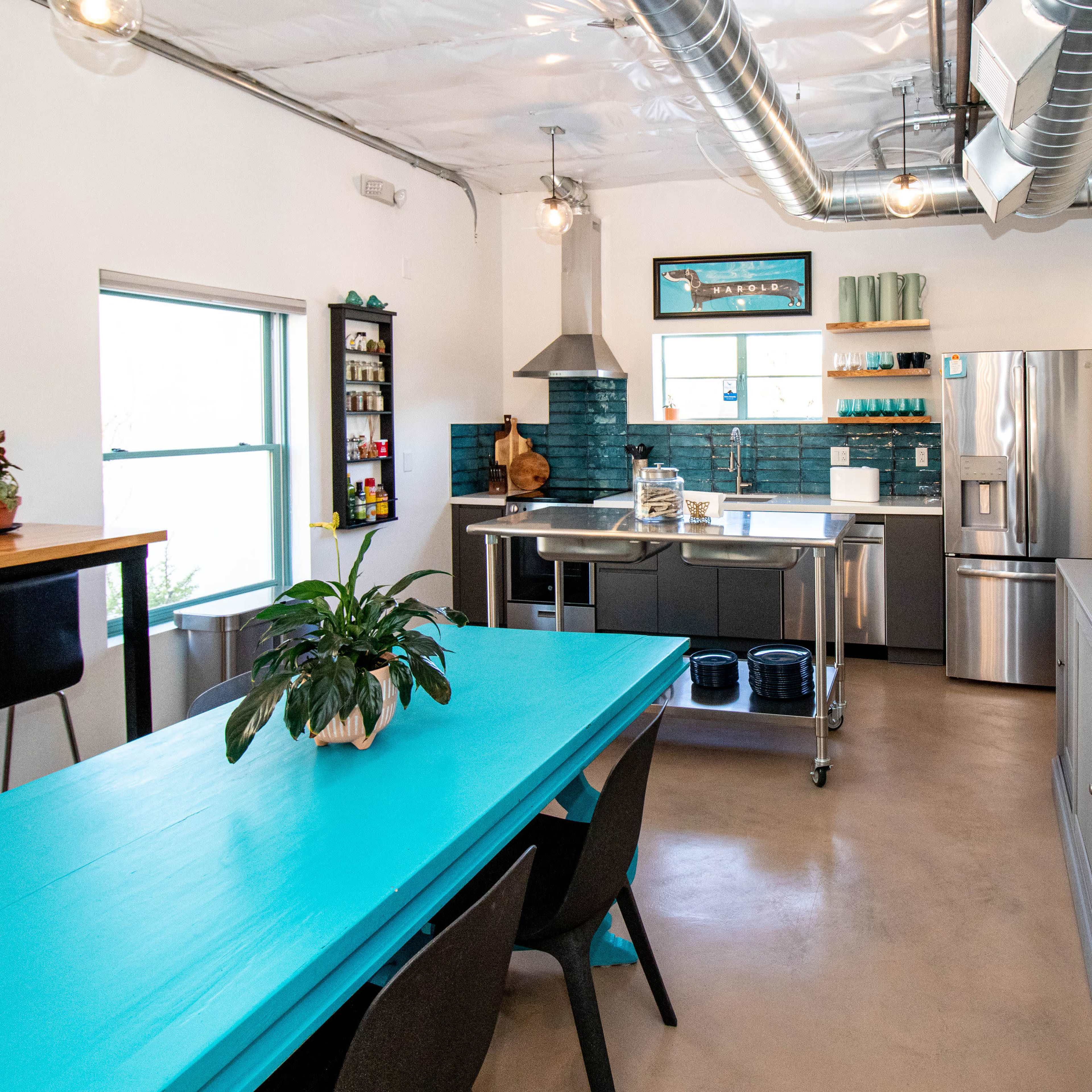 The kitchen features a turquoise dining table, stainless steel appliances, and an open layout with natural light.