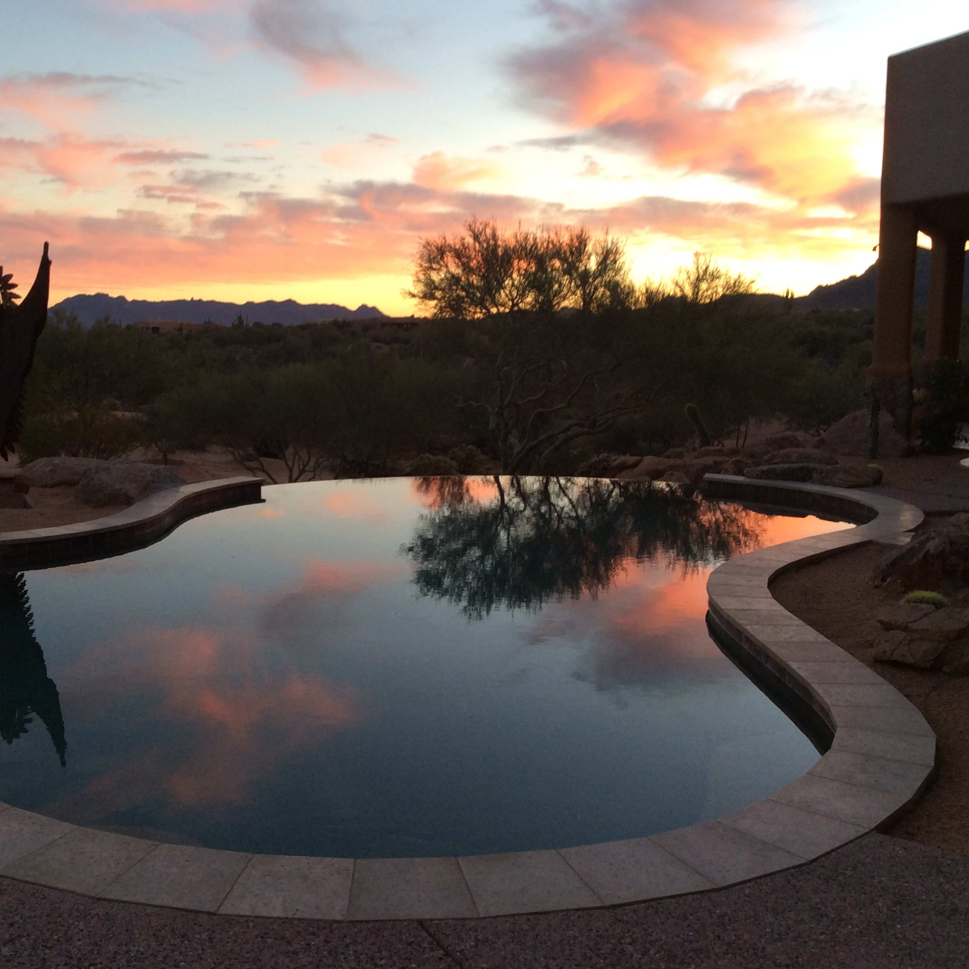 A serene swimming pool reflecting the colorful sunset sky, surrounded by desert landscaping and distant mountains.
