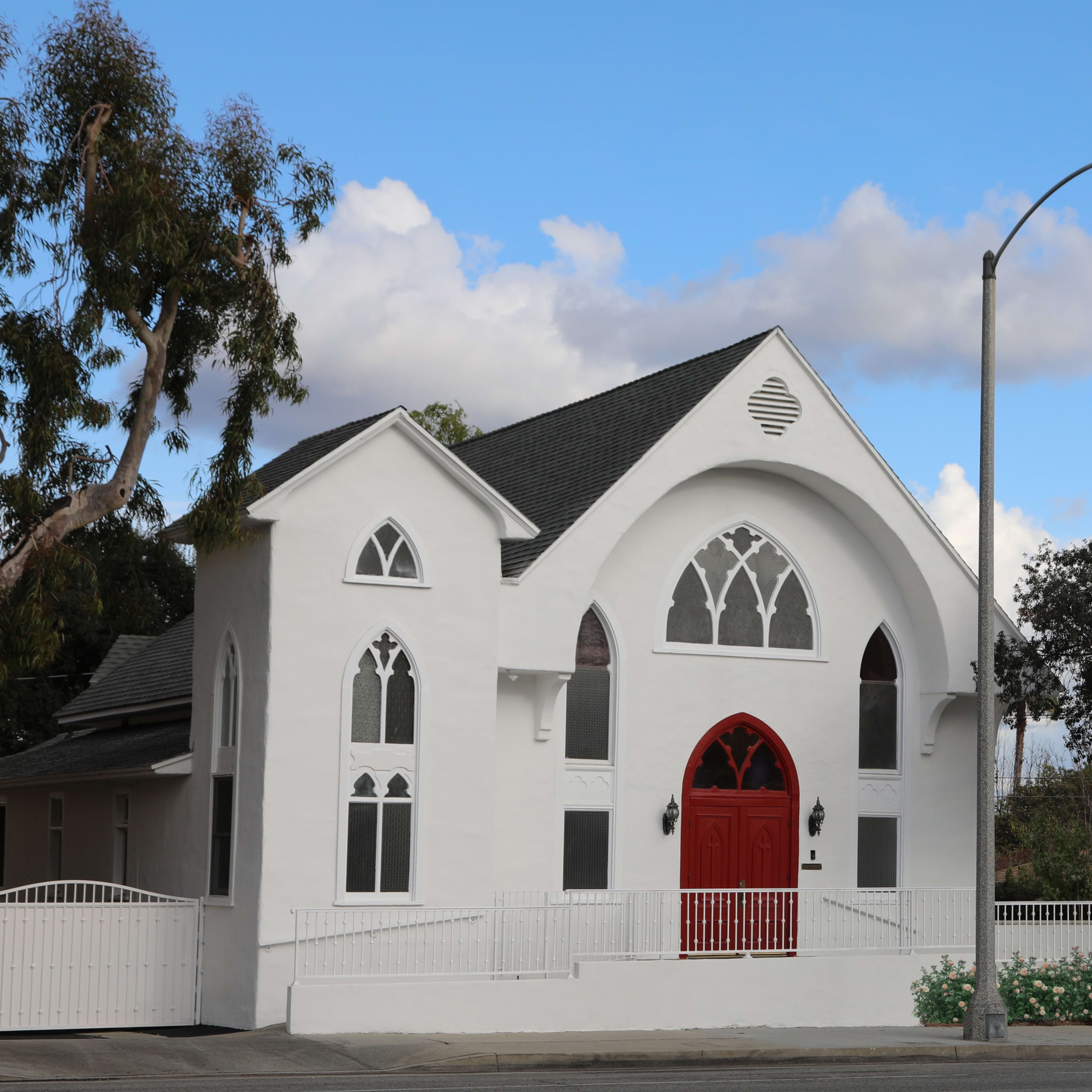 A white building with arched windows and a red door stands on a quiet street under a blue sky with scattered clouds.