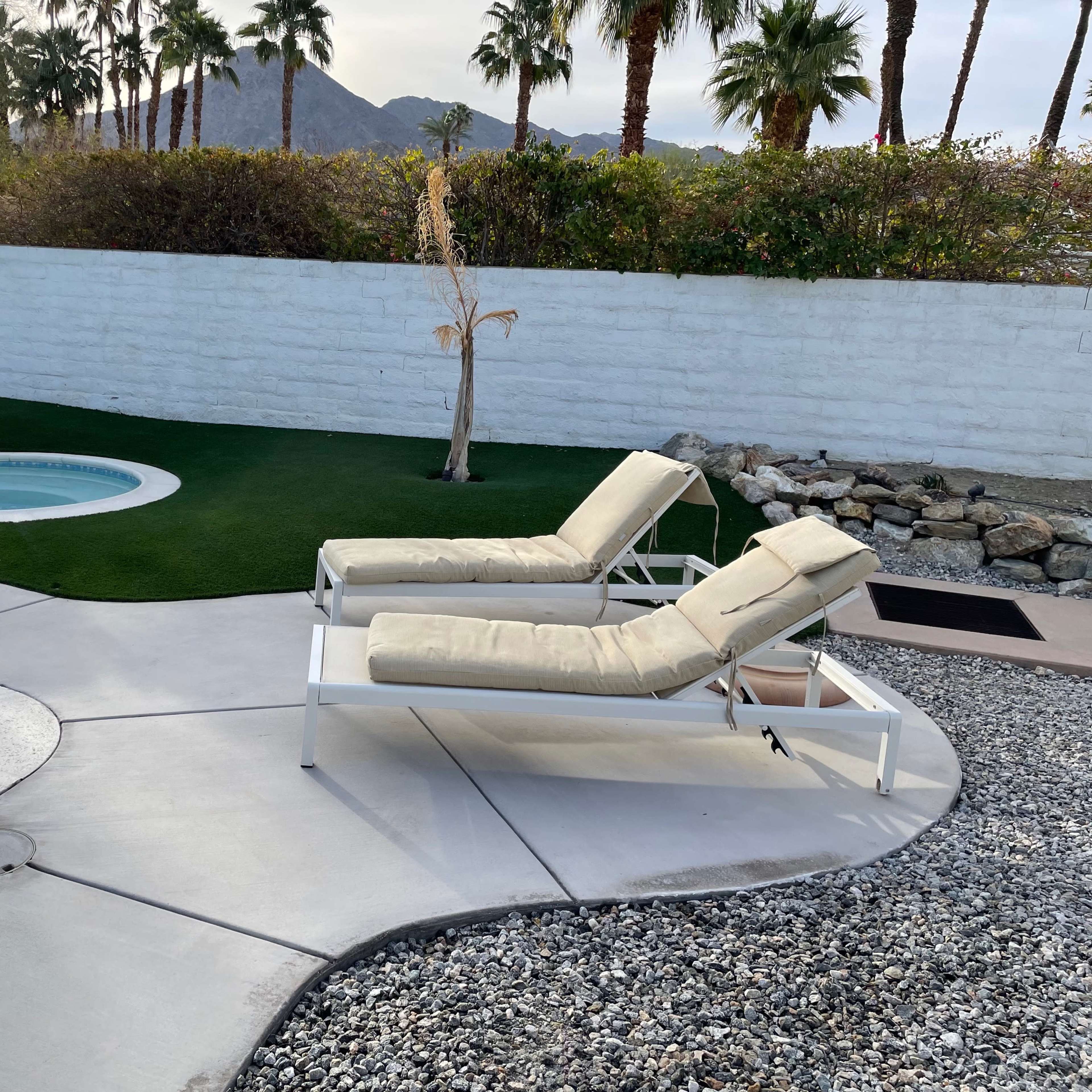 Two adjustable lounge chairs beside a swimming pool and a round hot tub, set against a backdrop of palm trees and a low white wall.