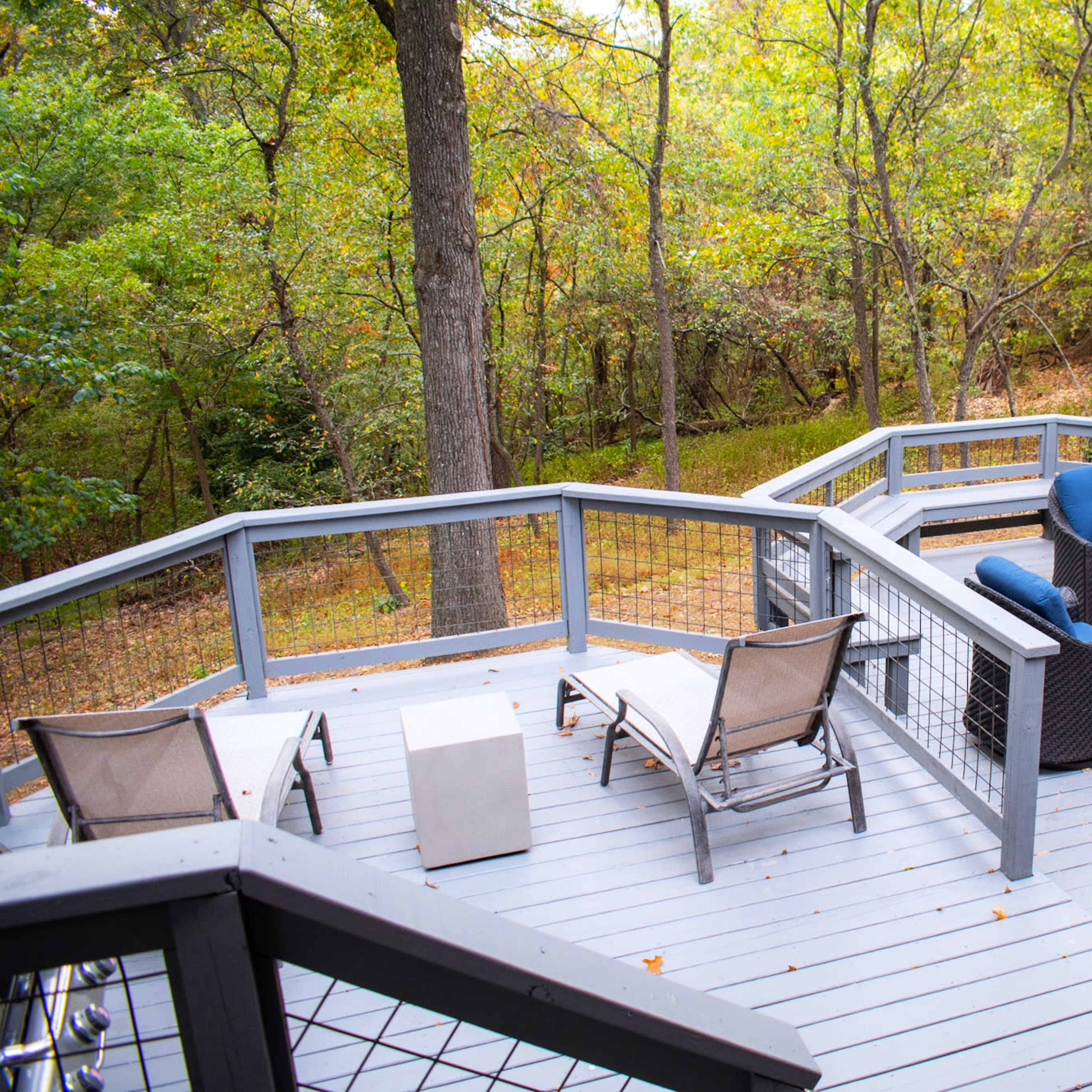A wooden deck features a set of blue cushioned chairs and a table with a fire pit, surrounded by trees with autumn foliage.