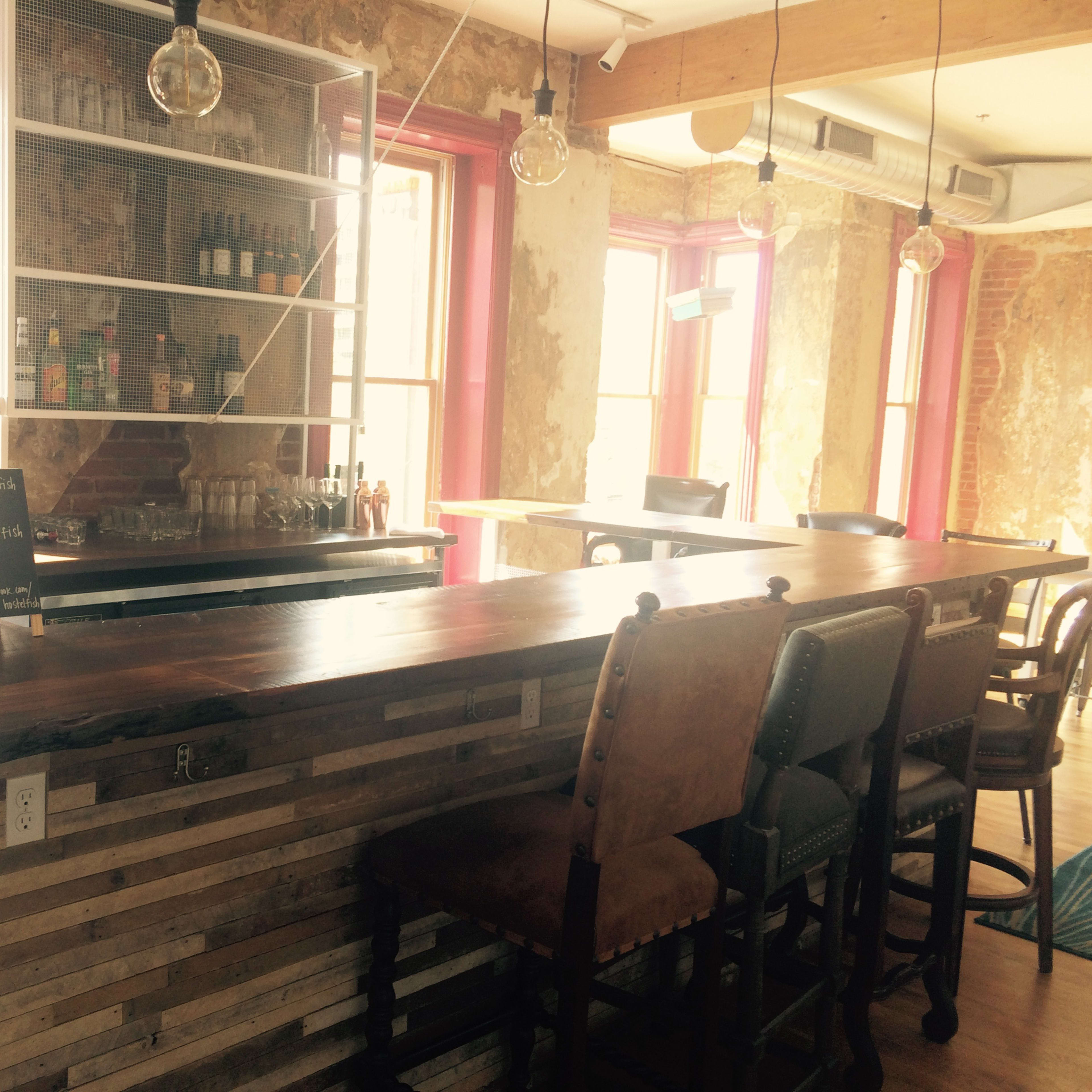 A rustic bar area with a wooden counter, several high-backed chairs, and a display of bottles in a well-lit interior with large windows.