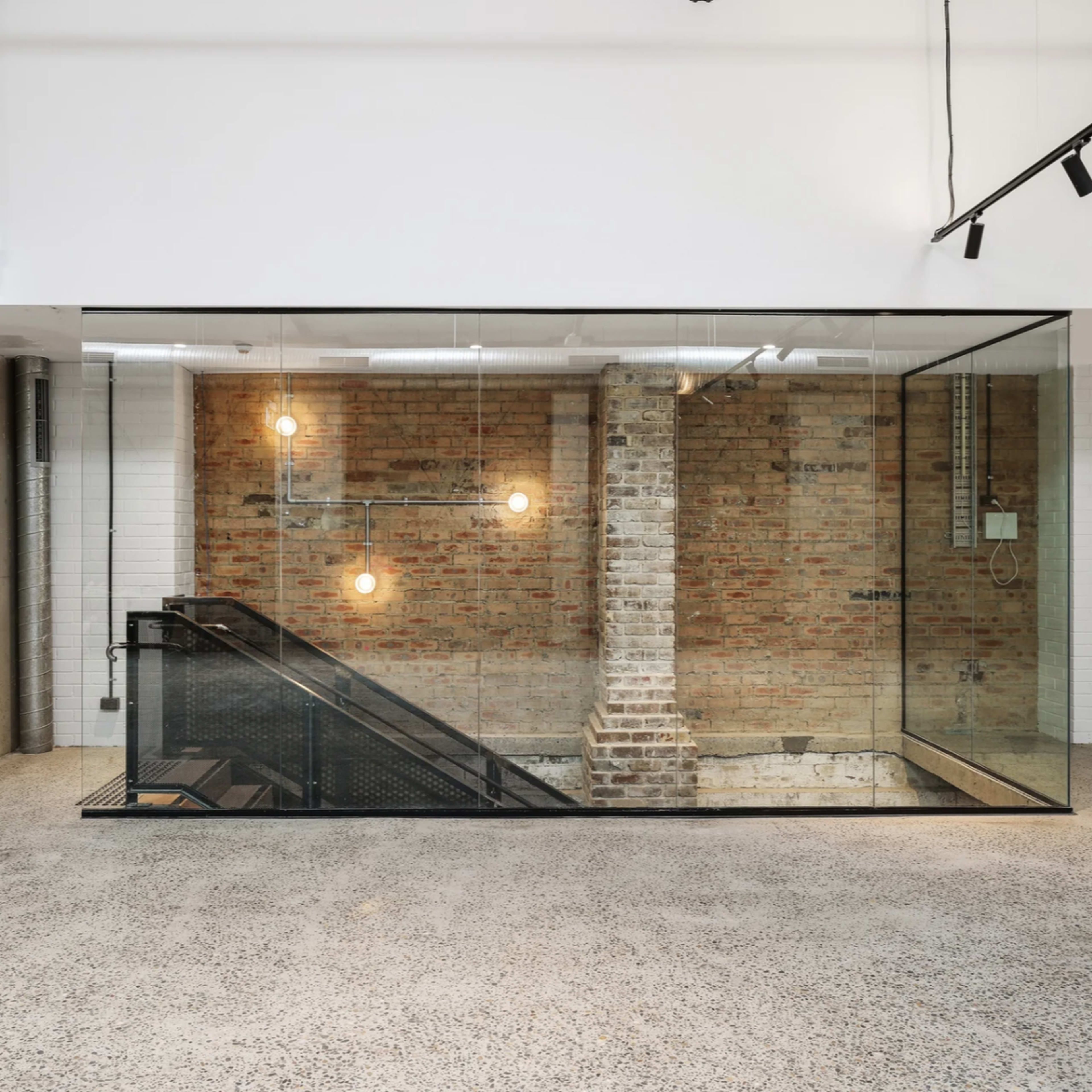 A modern interior space featuring a glass-walled staircase that leads to a lower level, with exposed brick walls and overhead lighting.