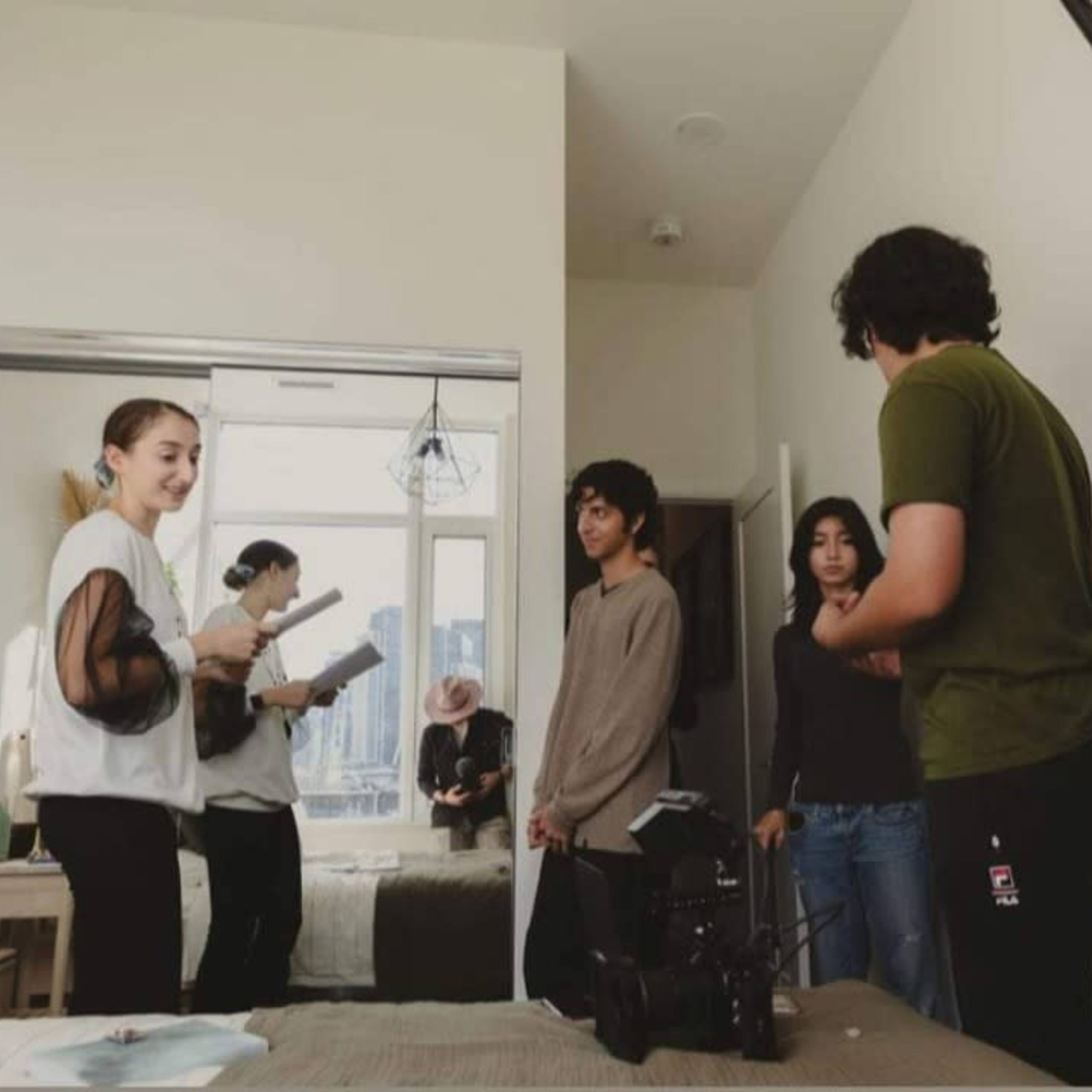 A group of five individuals is gathered in a bedroom, with two holding scripts and a camera, while others adjust equipment.