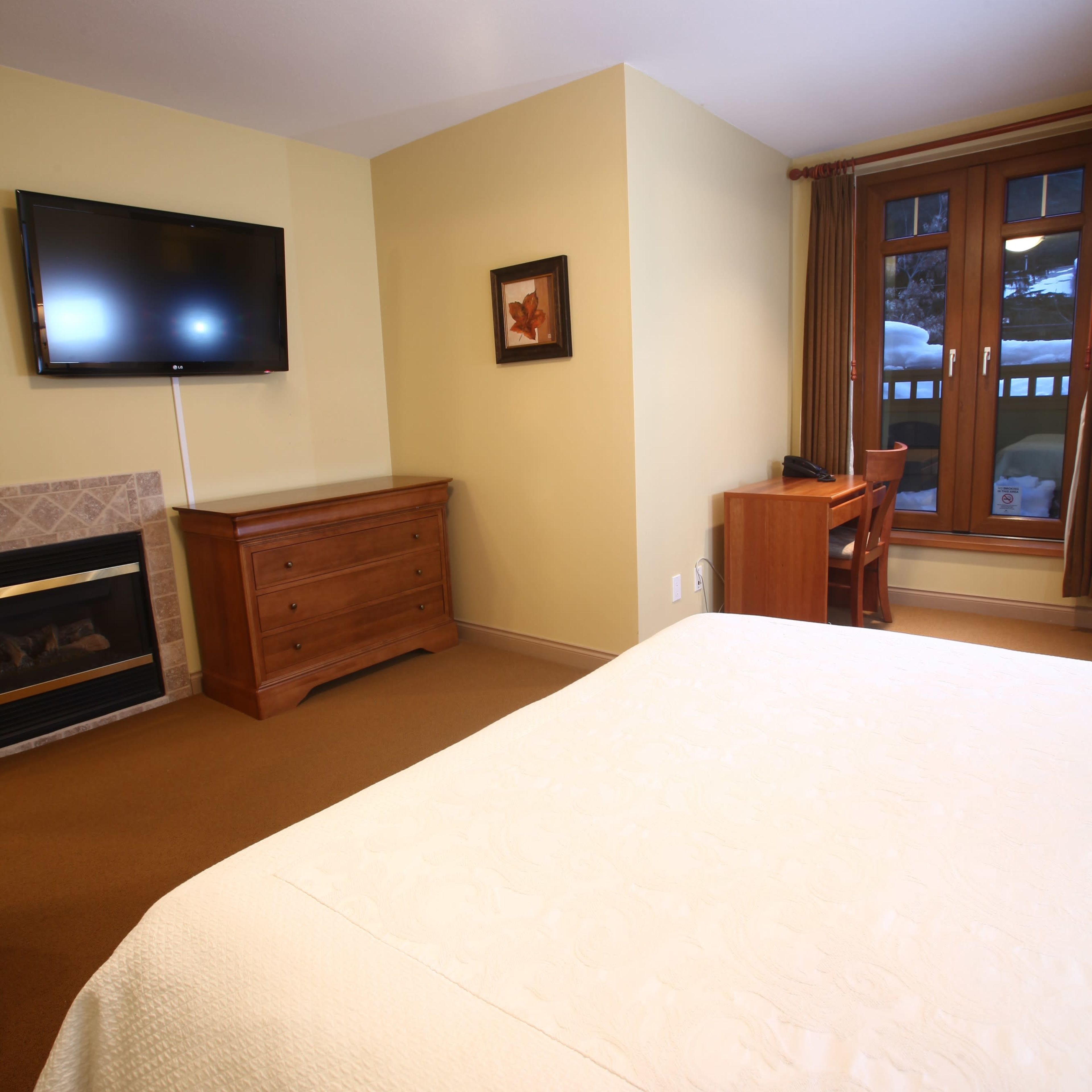 A hotel room featuring a bed, a wooden dresser, a television mounted on the wall, and a window with a desk and chair nearby.