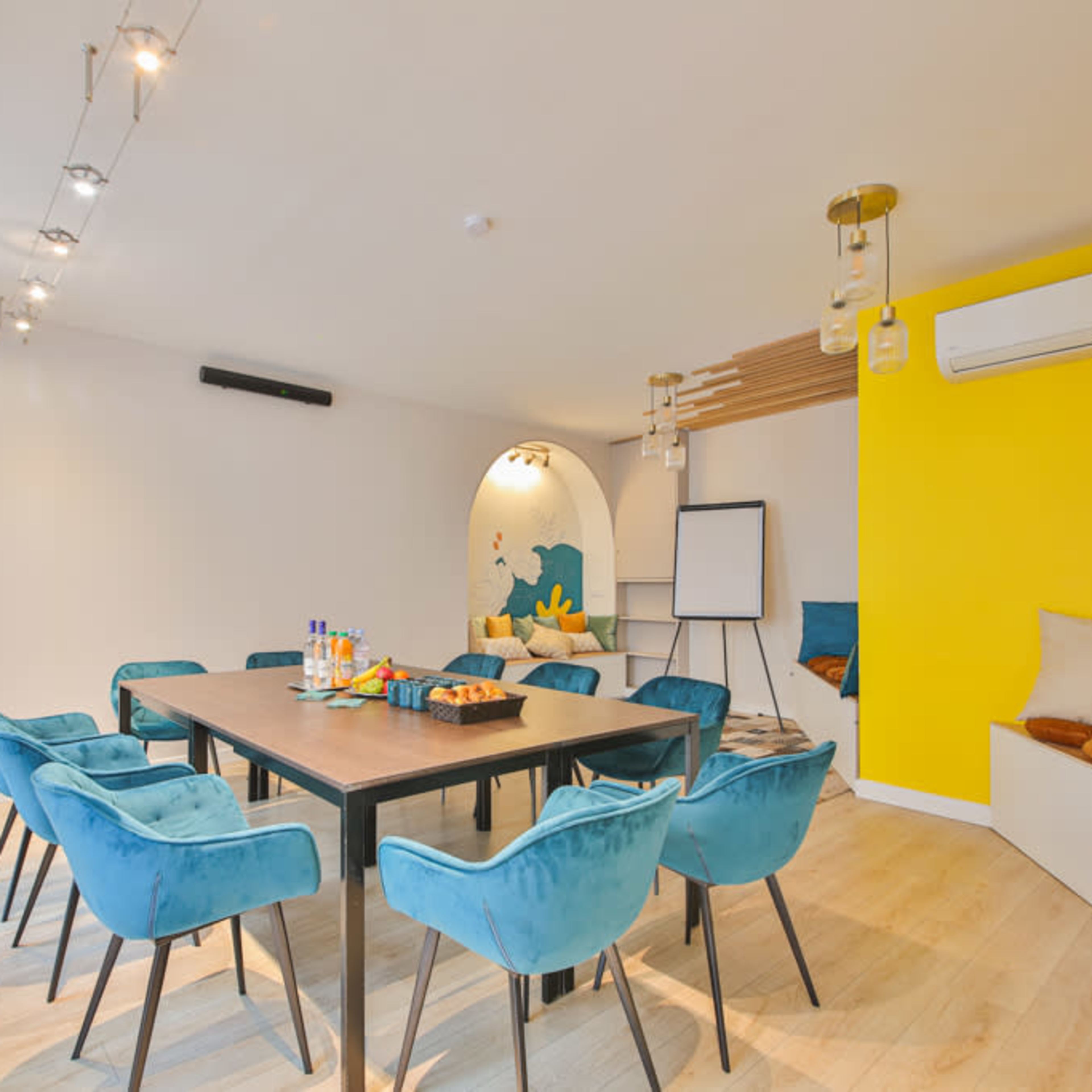 A modern meeting room features a large table surrounded by blue chairs, a whiteboard, and bright yellow accents on the wall.