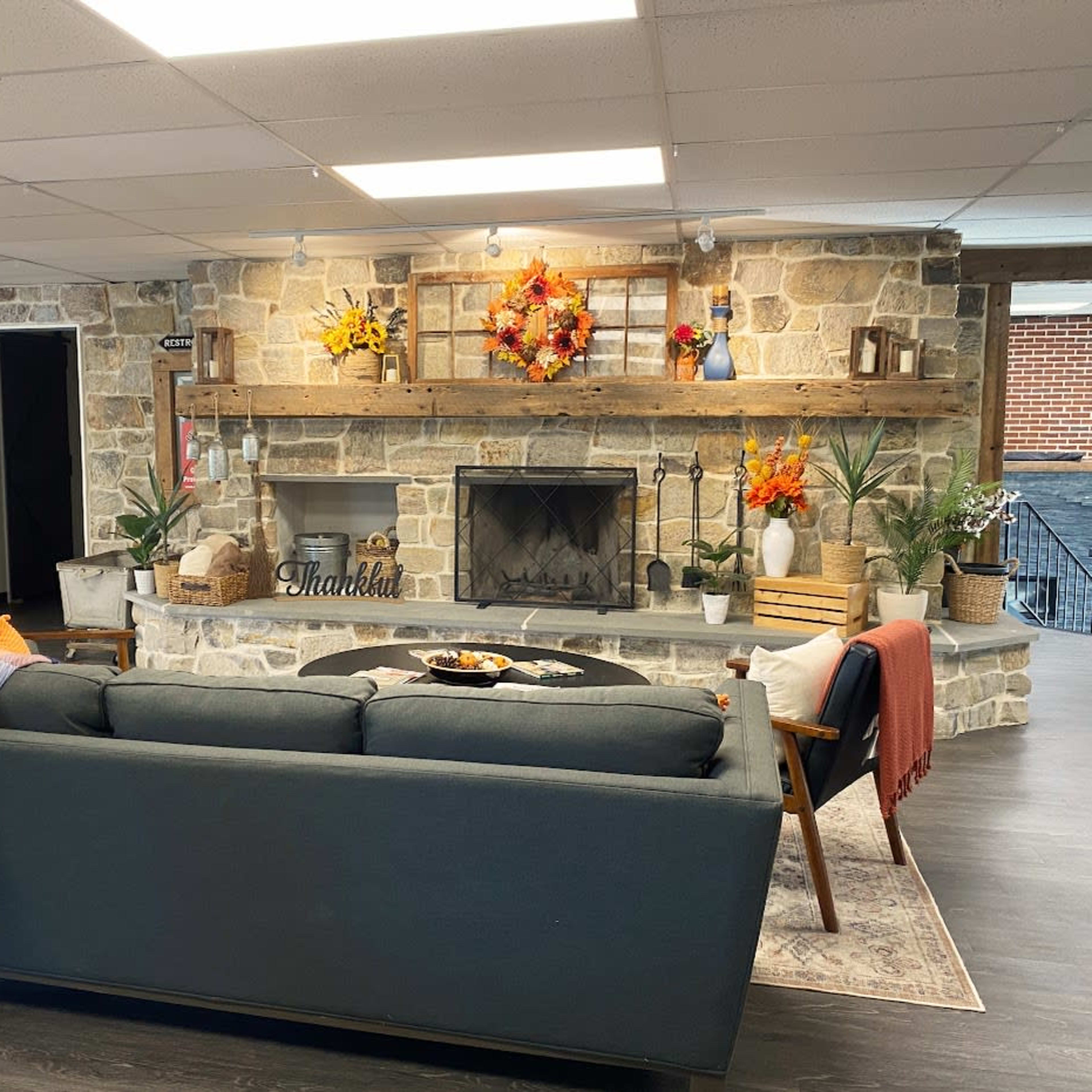 A cozy living area with a stone fireplace, a dark sofa, a patterned blanket draped over the back, and decorative plants and flowers arranged on the mantel.