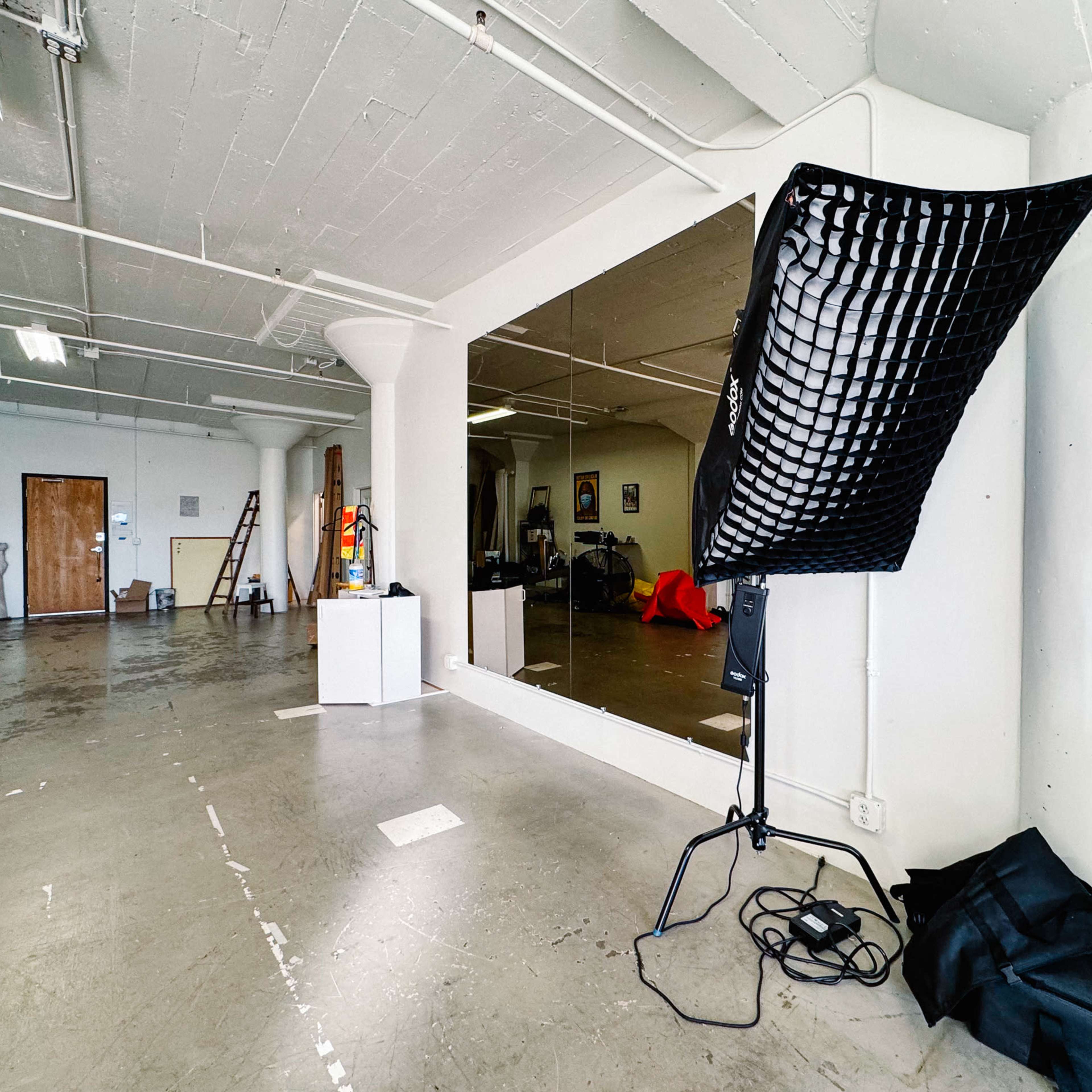 A spacious, unfinished studio with a large mirror, lighting equipment, and scattered furniture.