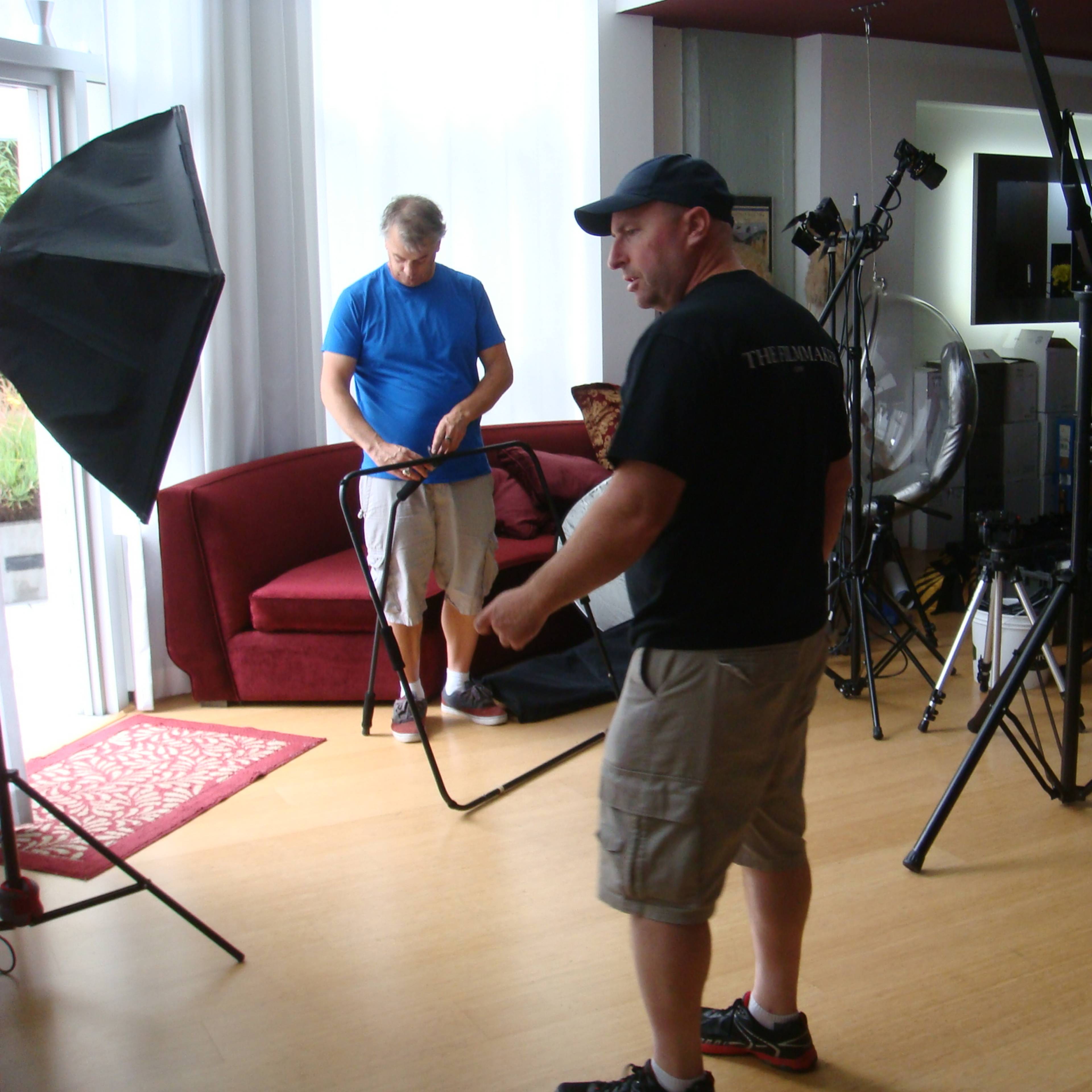 Two men set up equipment in a well-lit studio with a red couch and various lighting stands in the background.