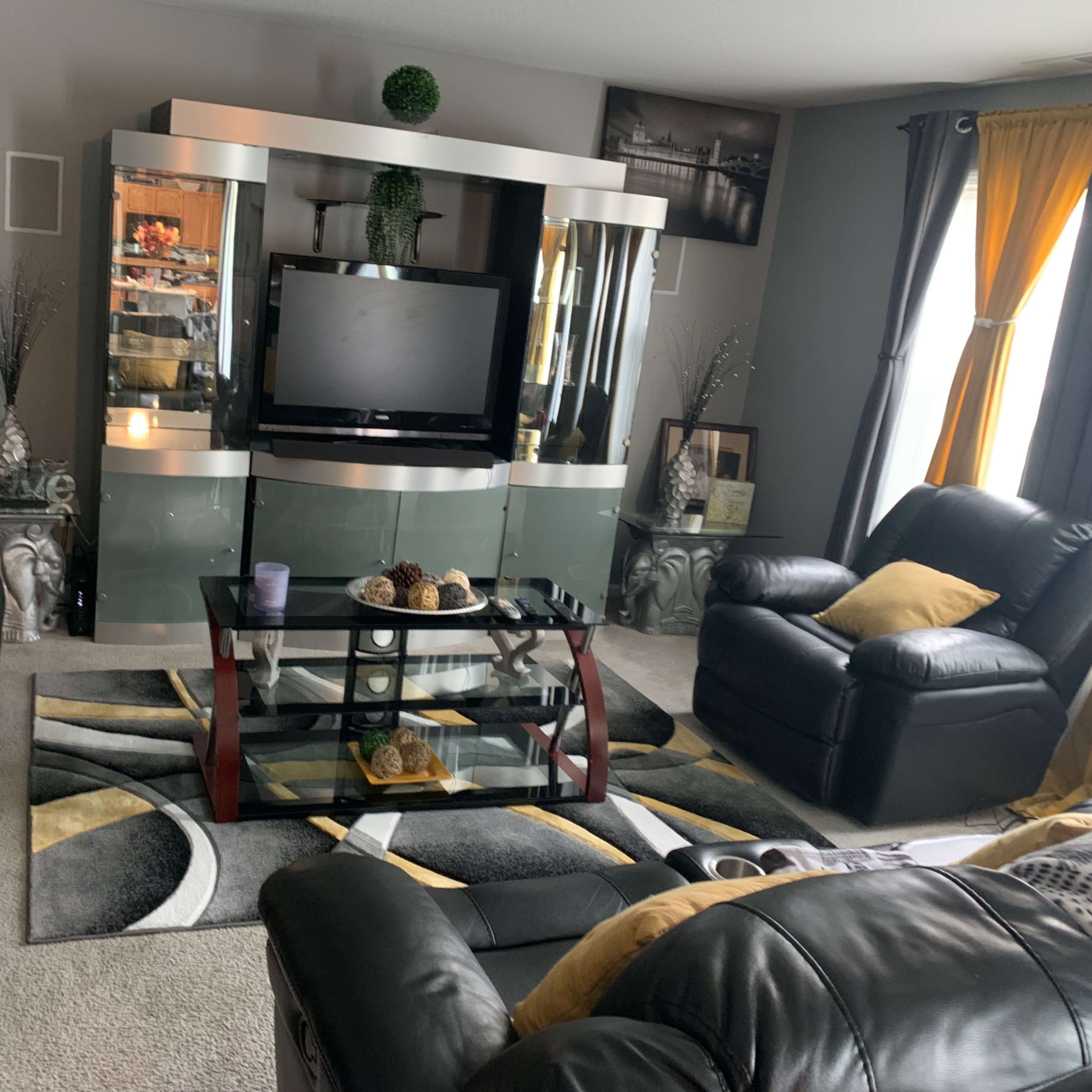 The living room features black leather furniture, a modern glass entertainment center with a television, and decorative accents on a patterned area rug.