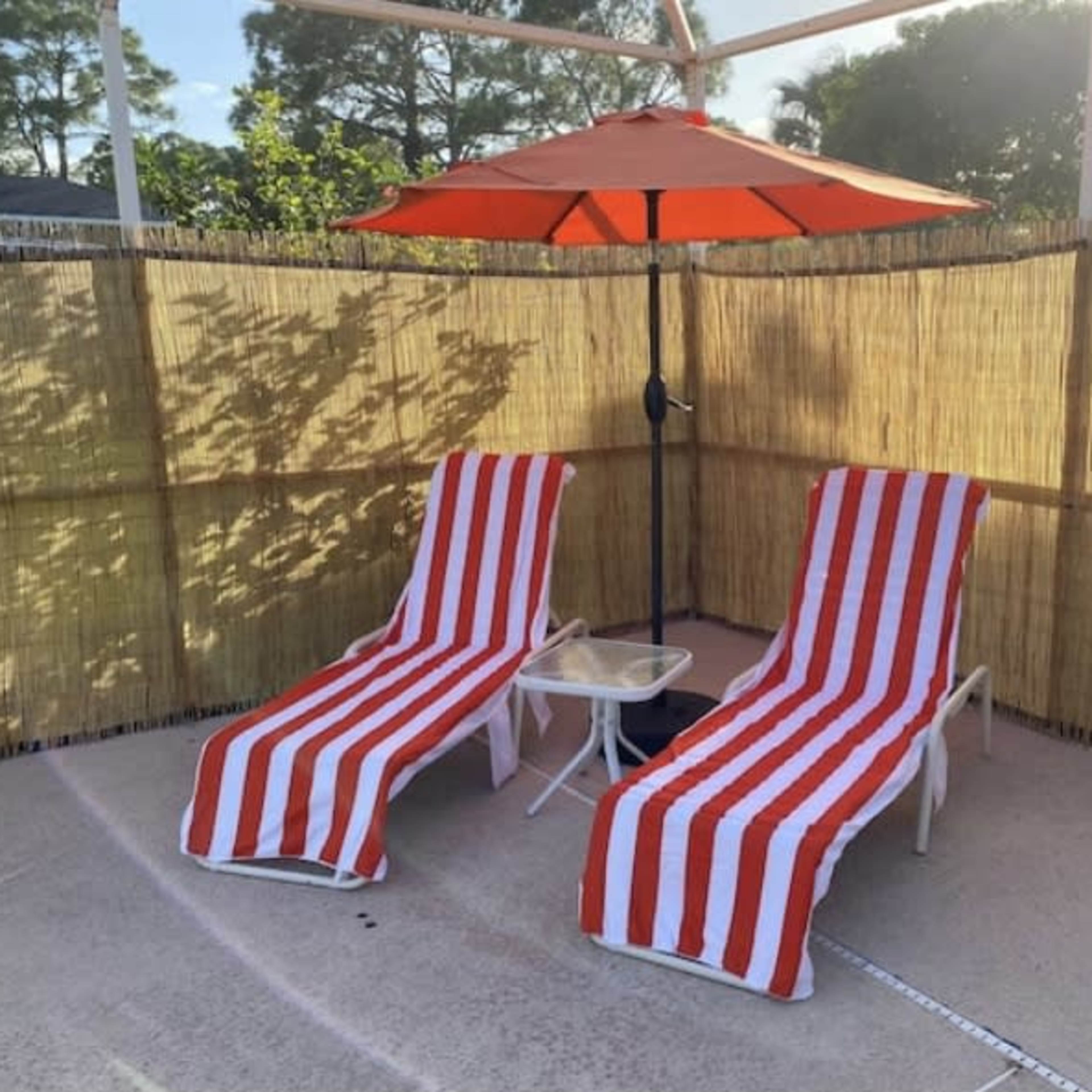 Two striped lounge chairs with a small table are positioned under a red umbrella, surrounded by a bamboo screen.