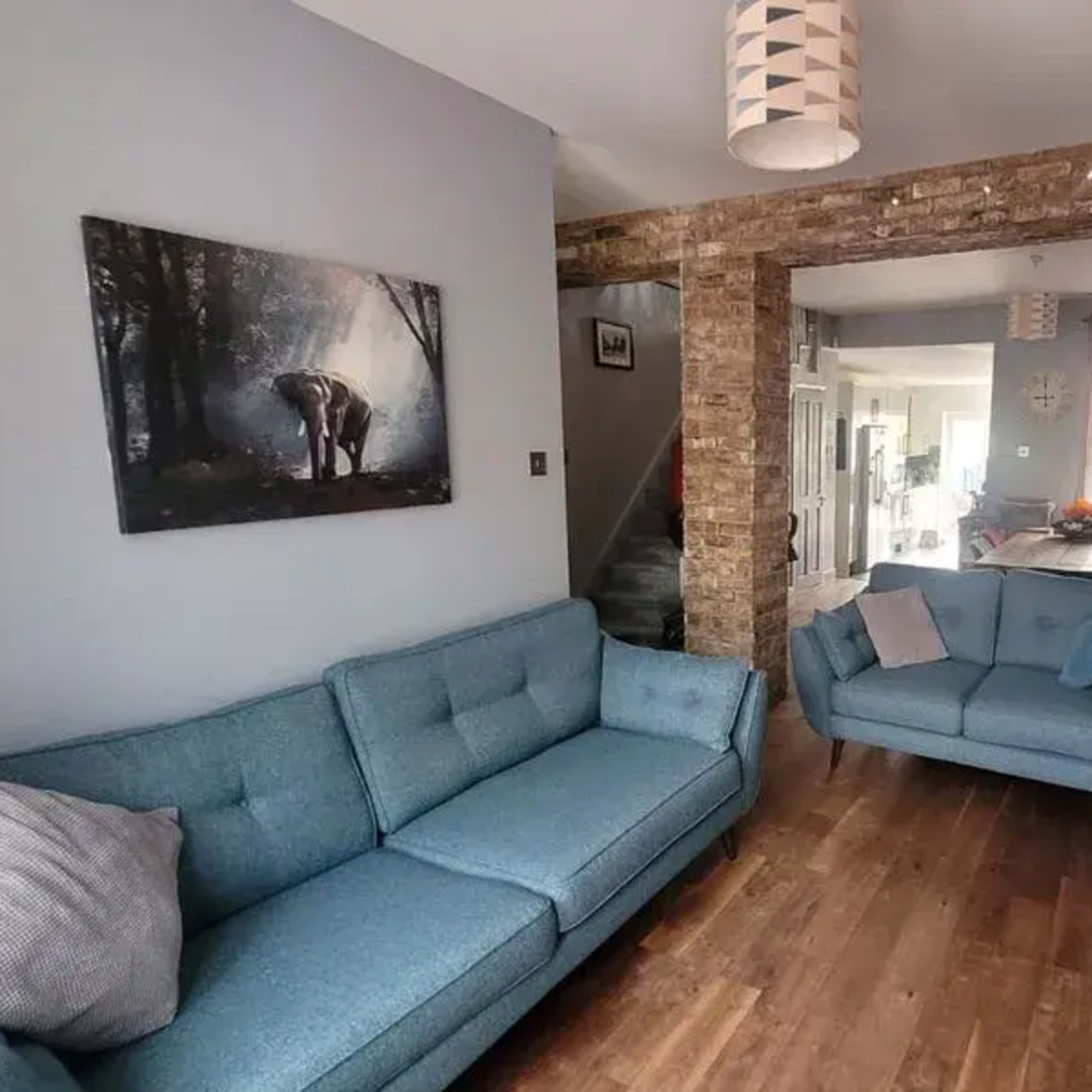 The room features two blue sofas, a stone wall, and a television mounted on the wall, with an open space leading to a dining area visible in the background.