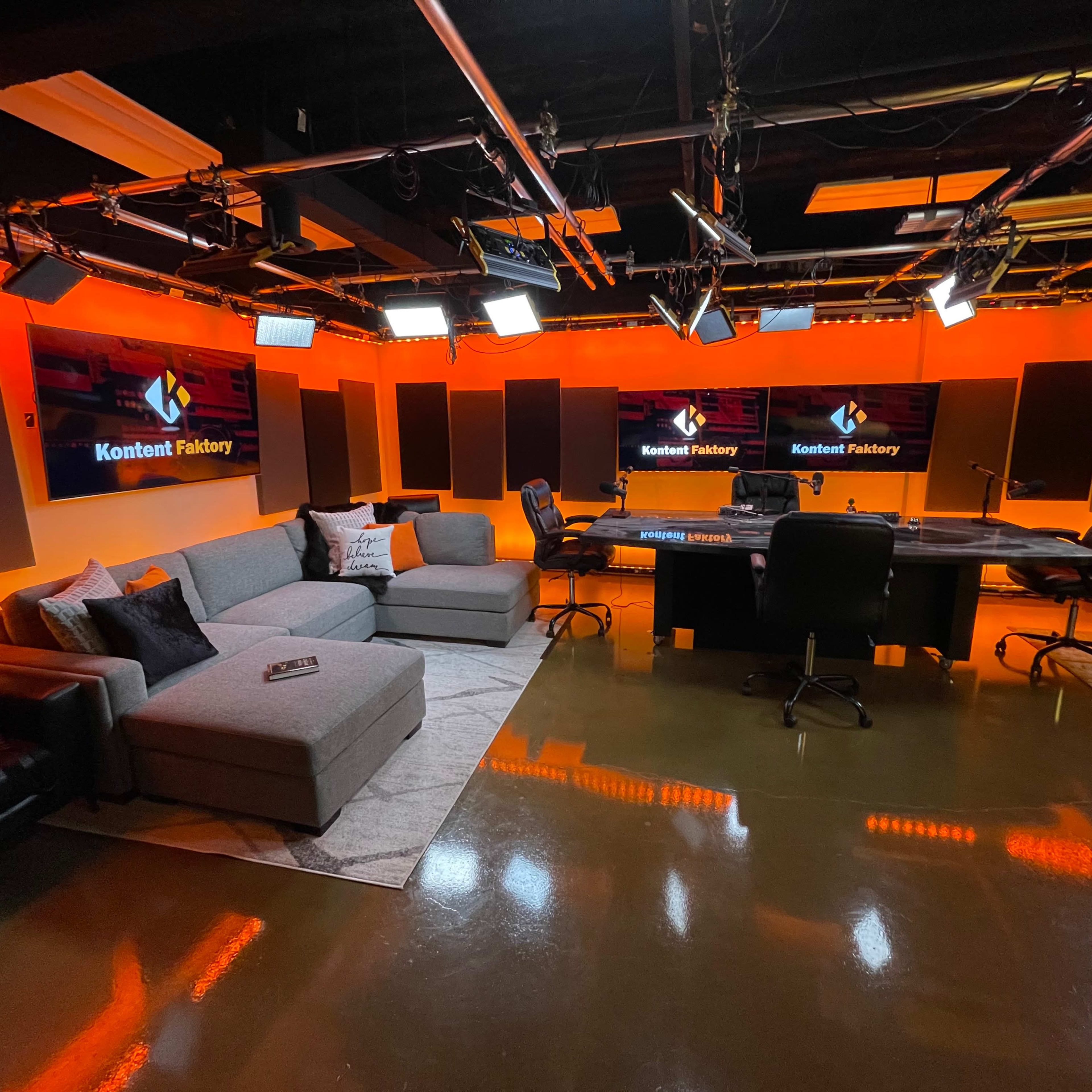A studio setting with orange-lit walls, plush seating, a large conference table, and multiple screens displaying the text "Kontent Factory."