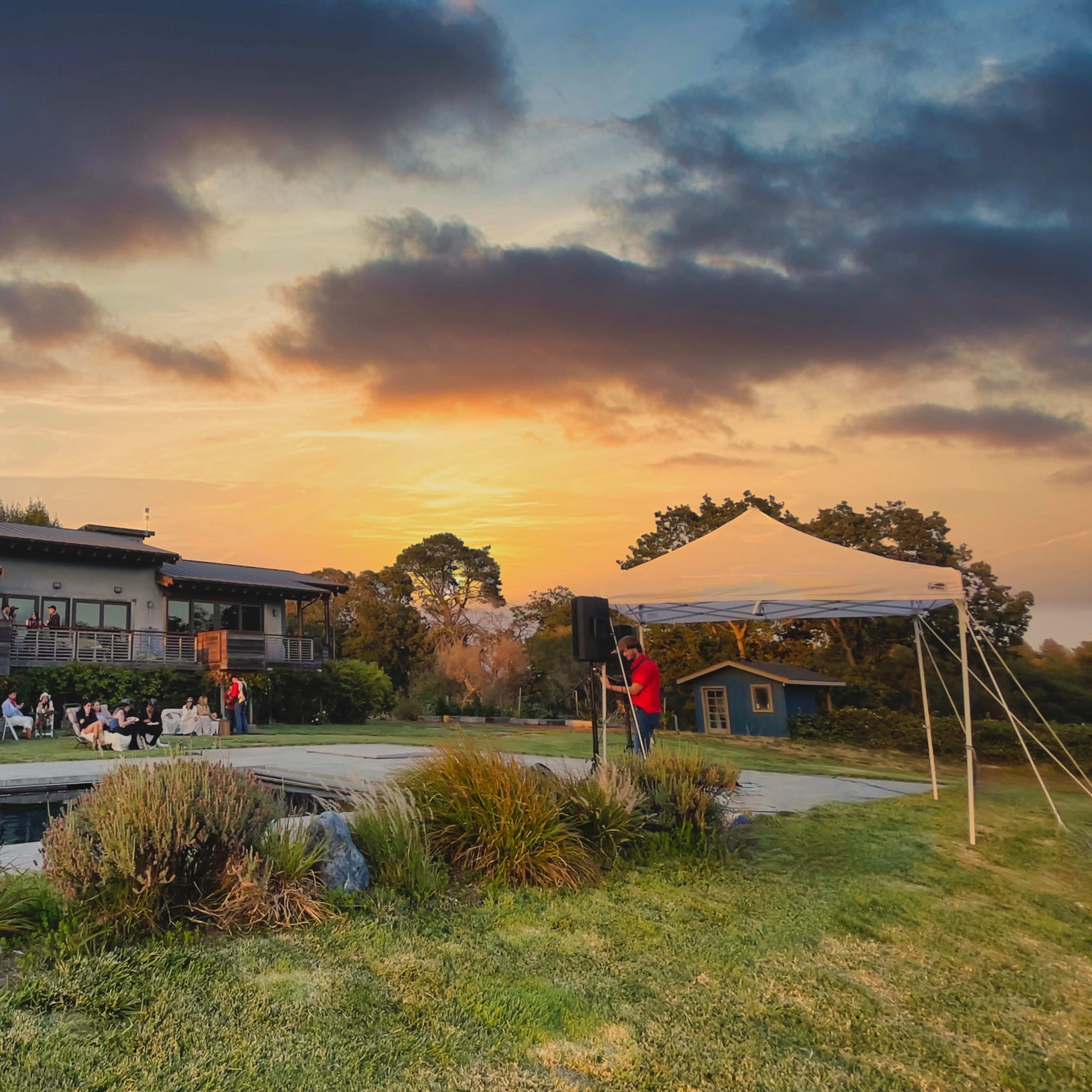 A large gathering of people sits outside a building during sunset, with a tent set up nearby and a pond in the foreground.