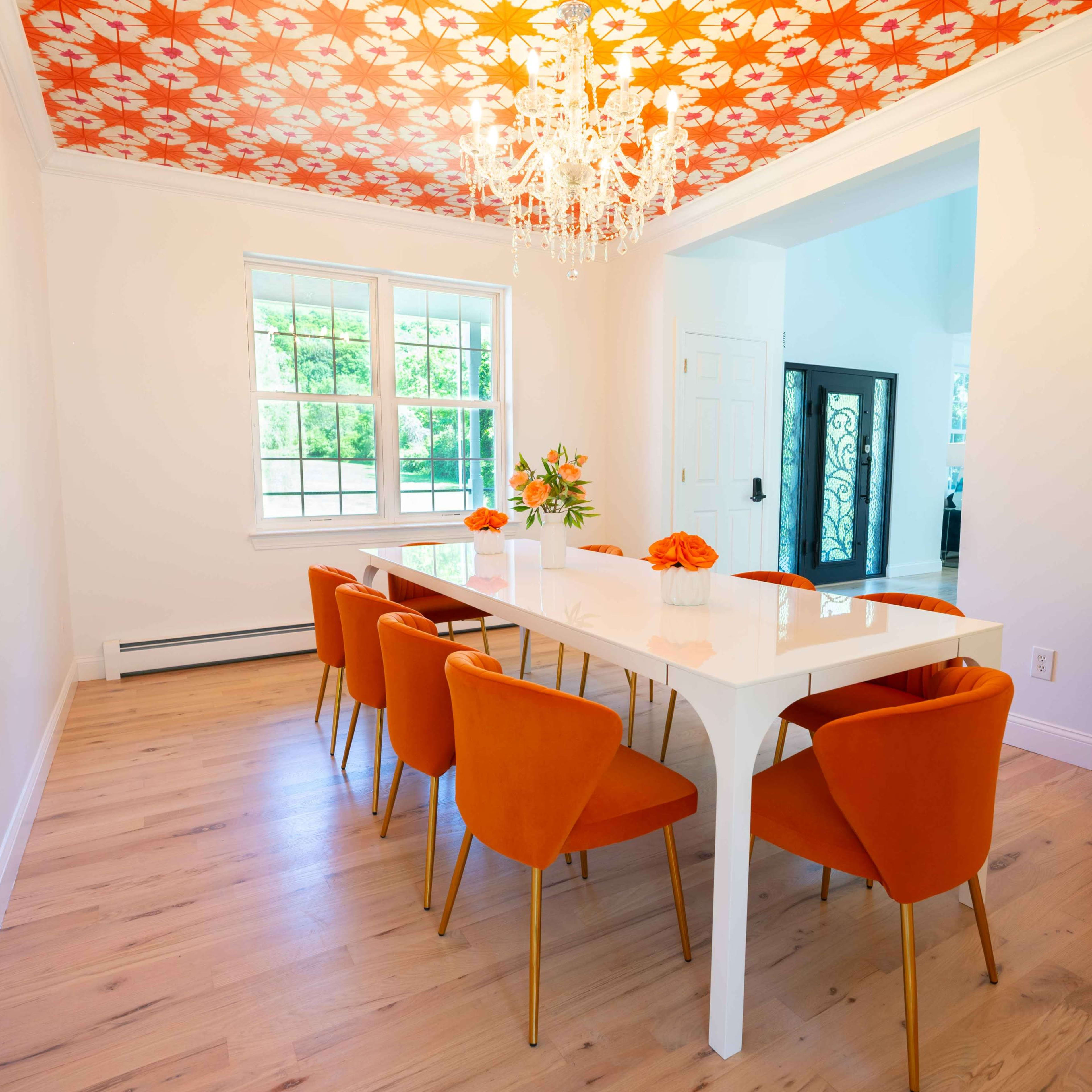 A modern dining room features a long white table surrounded by orange chairs, with a floral-patterned ceiling and a chandelier above.