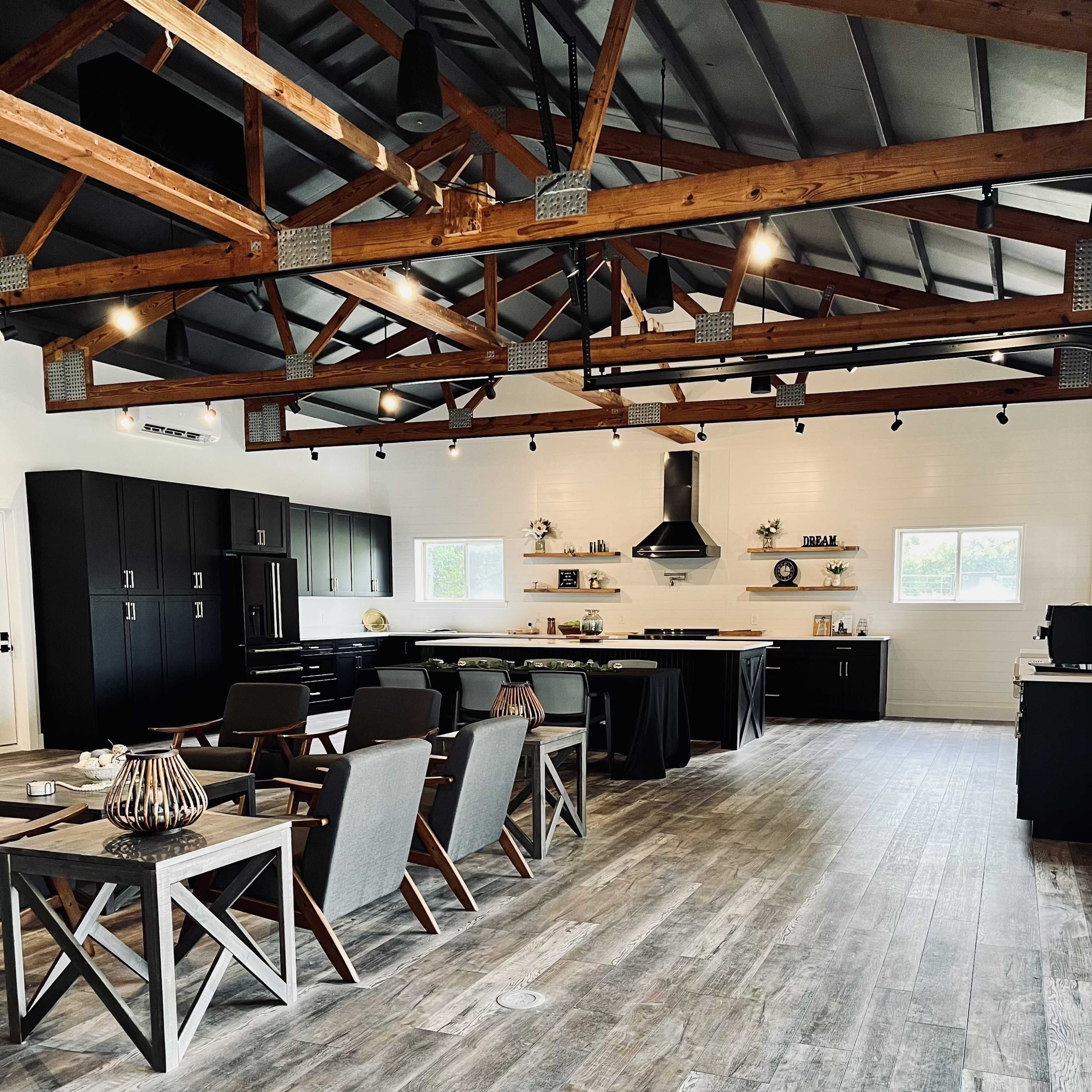 A modern kitchen and dining area with exposed wooden beams, black cabinetry, and a polished wooden floor, featuring seating arrangements and kitchen appliances.