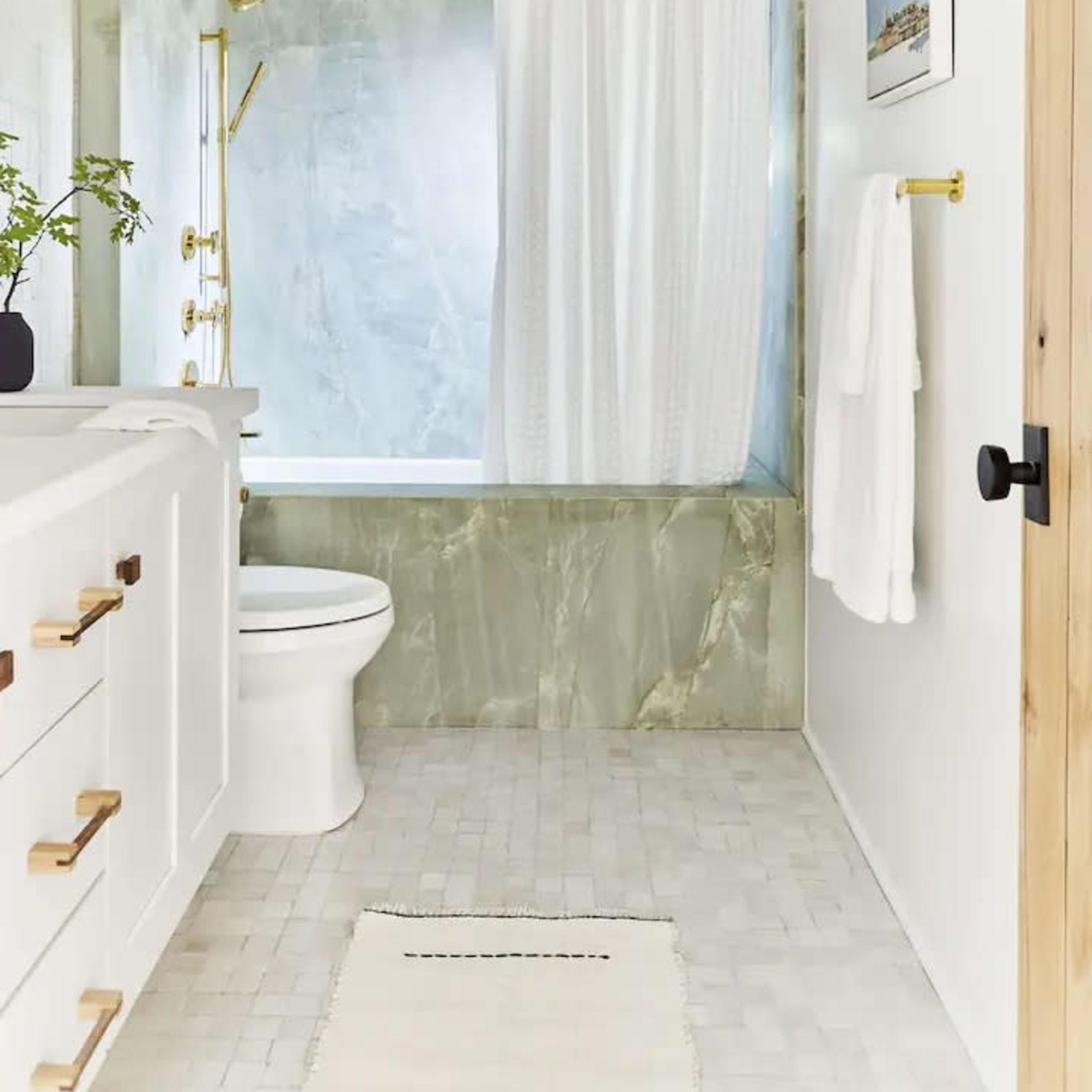 The modern bathroom features a white vanity with gold hardware, a green marble tub surround, and a shower curtain, alongside a white toilet and a light-colored rug.