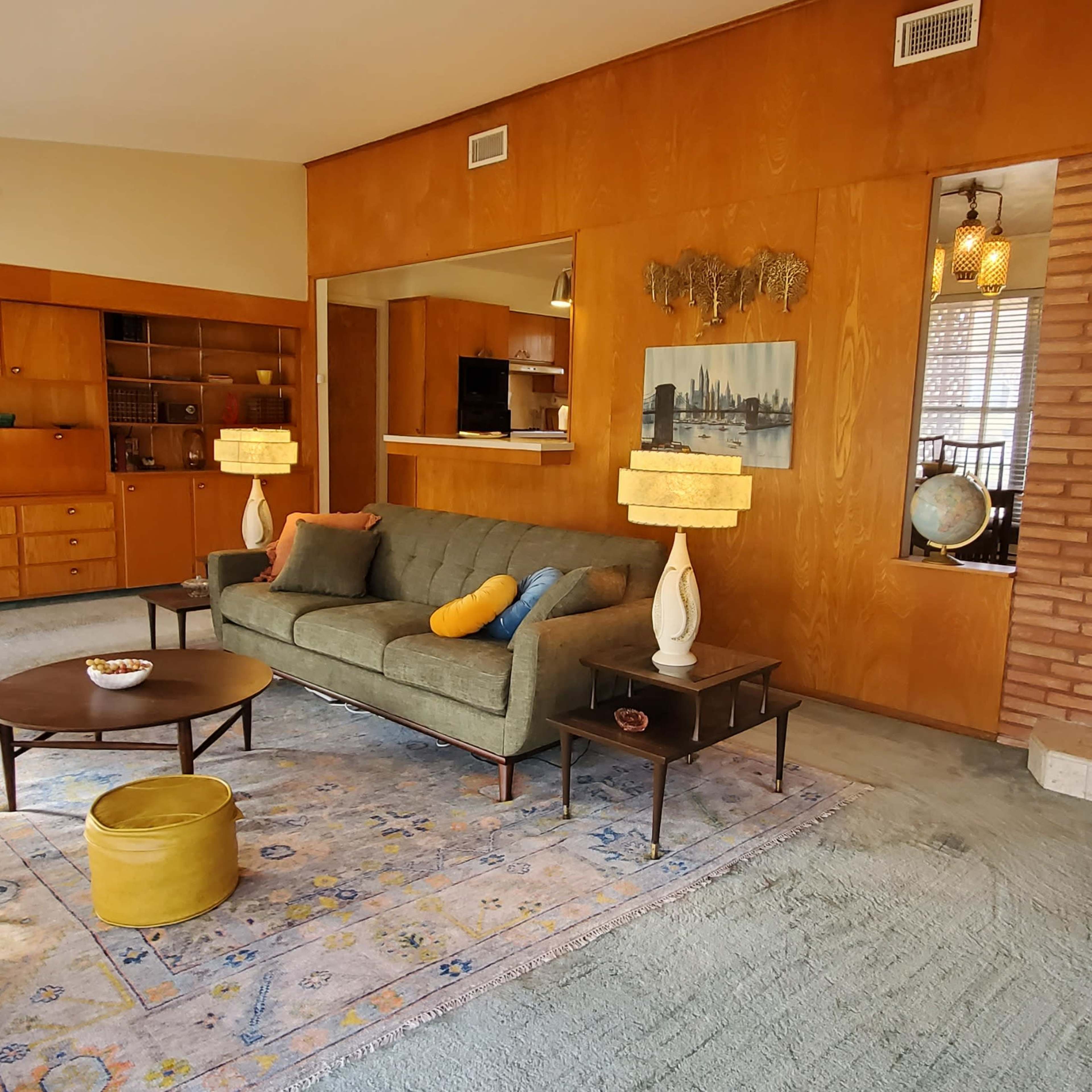 A mid-century modern living room featuring a green sofa, wooden paneling, a round coffee table, and decorative lamps, with a small staircase leading to a door.