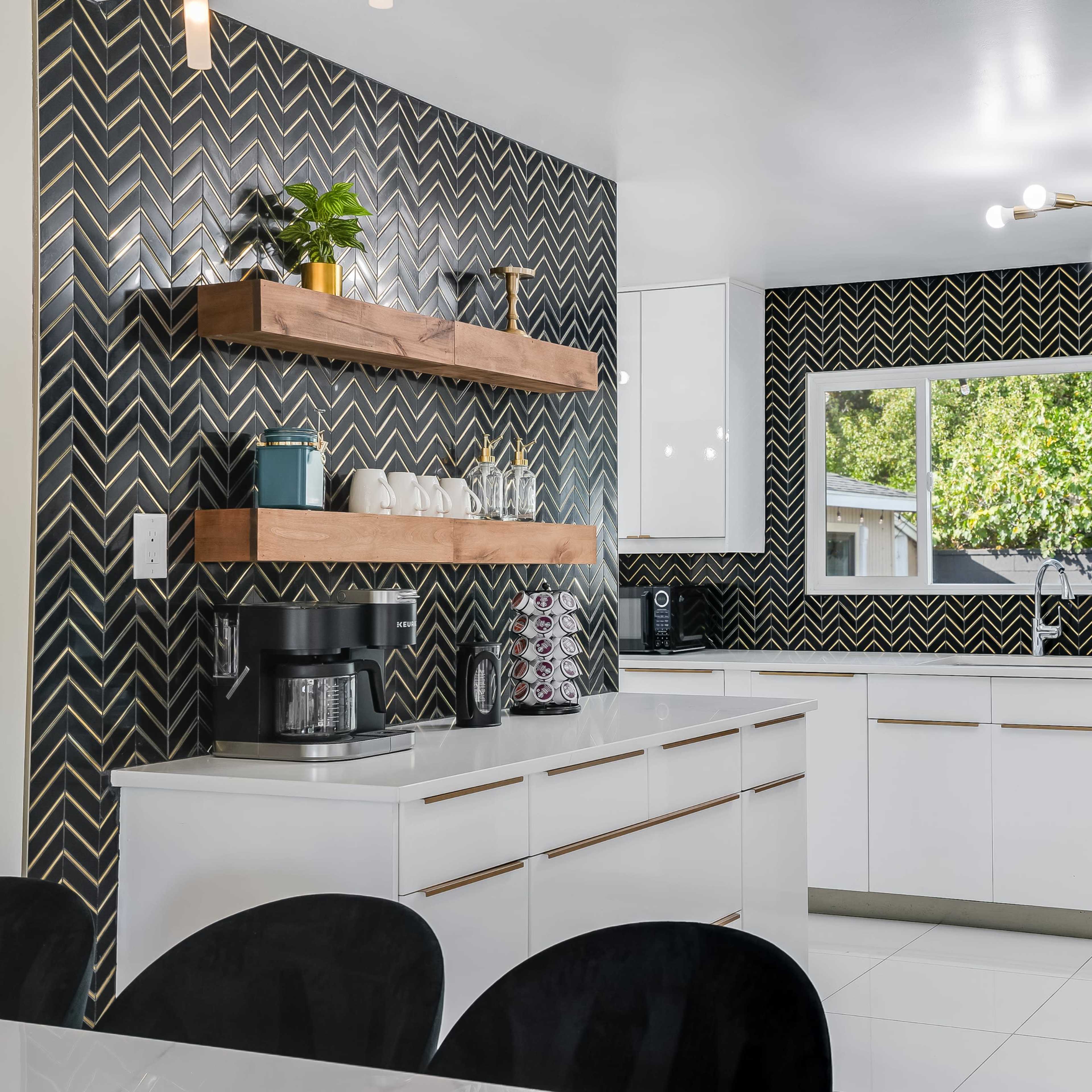 A modern kitchen features white cabinetry, black and gold patterned walls, wooden shelves, and a large window overlooking greenery.