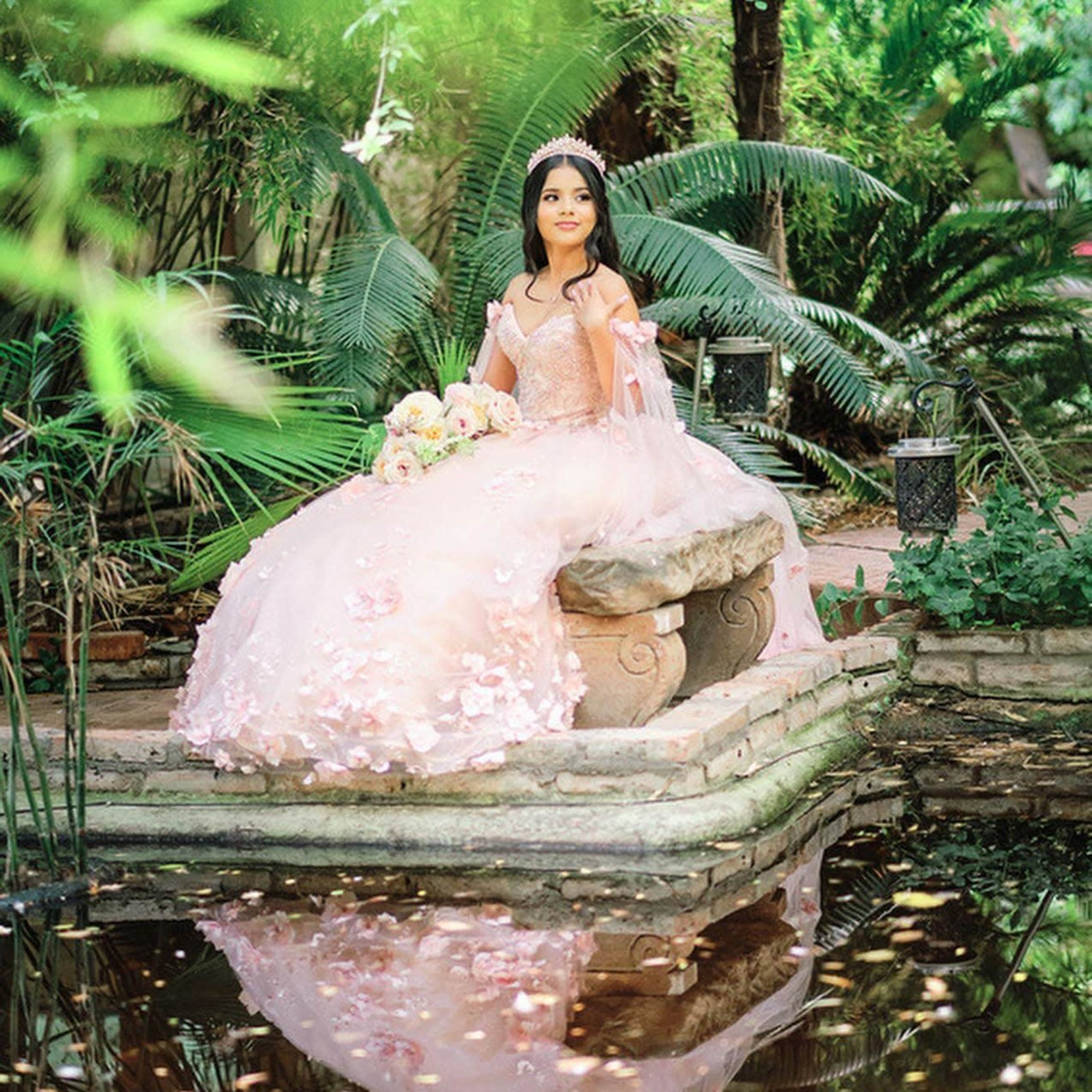 A young woman in a frilly pink gown holds a bouquet while sitting on a stone bench surrounded by greenery and reflecting water.