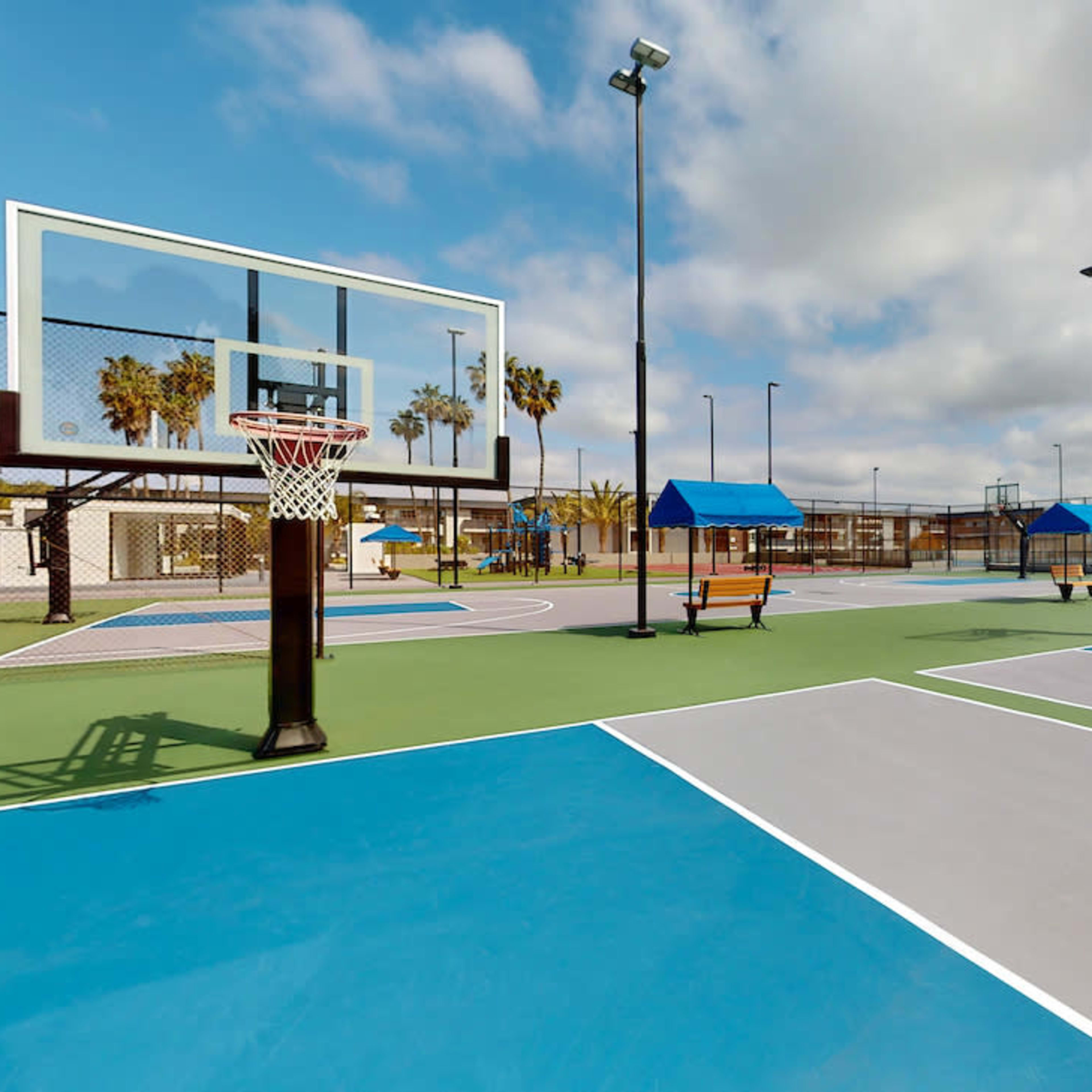 Several basketball courts with multiple hoops surrounded by palm trees under a partly cloudy sky.