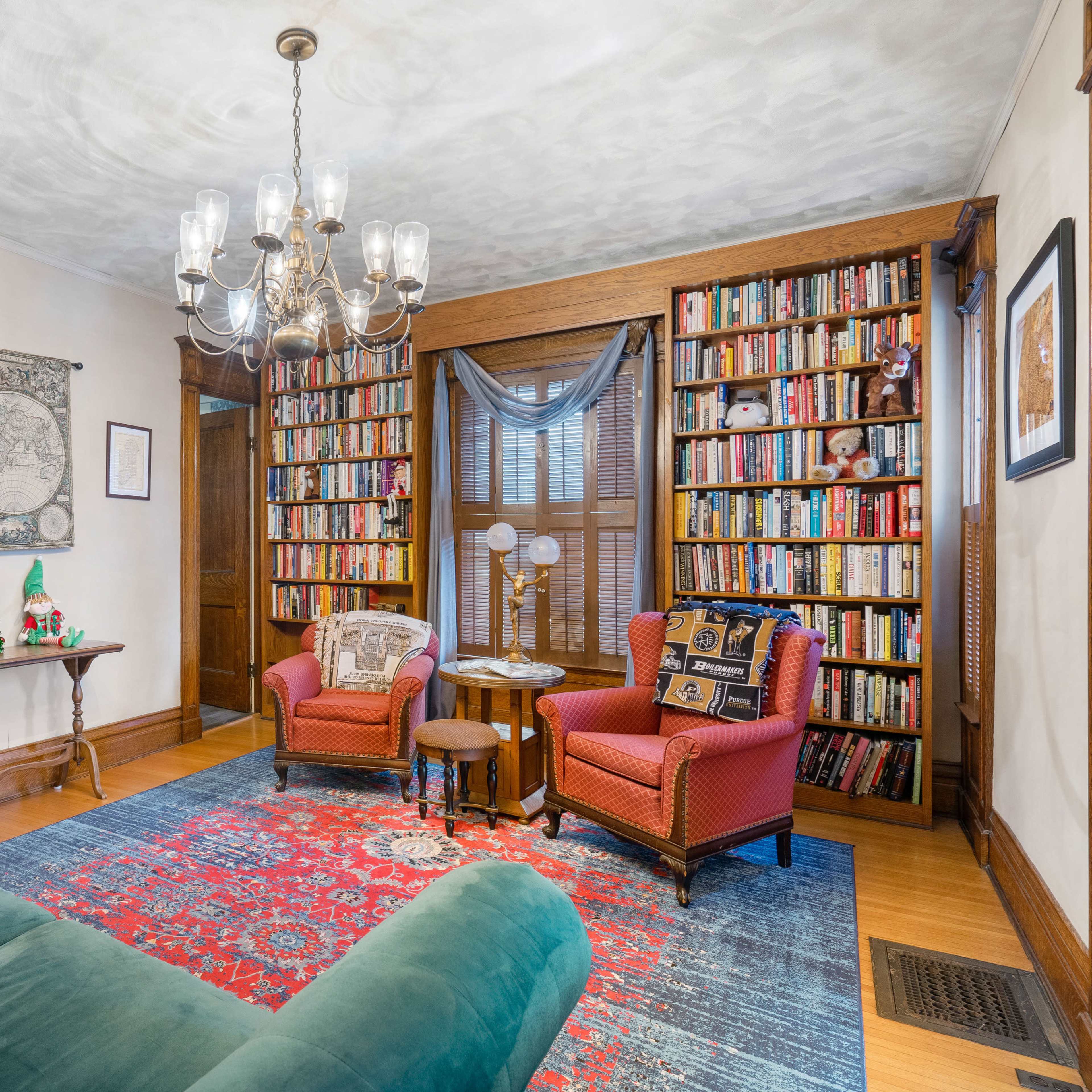 A cozy living room featuring a blue sofa, two red armchairs, a wooden bookshelf filled with books, and a chandelier overhead.