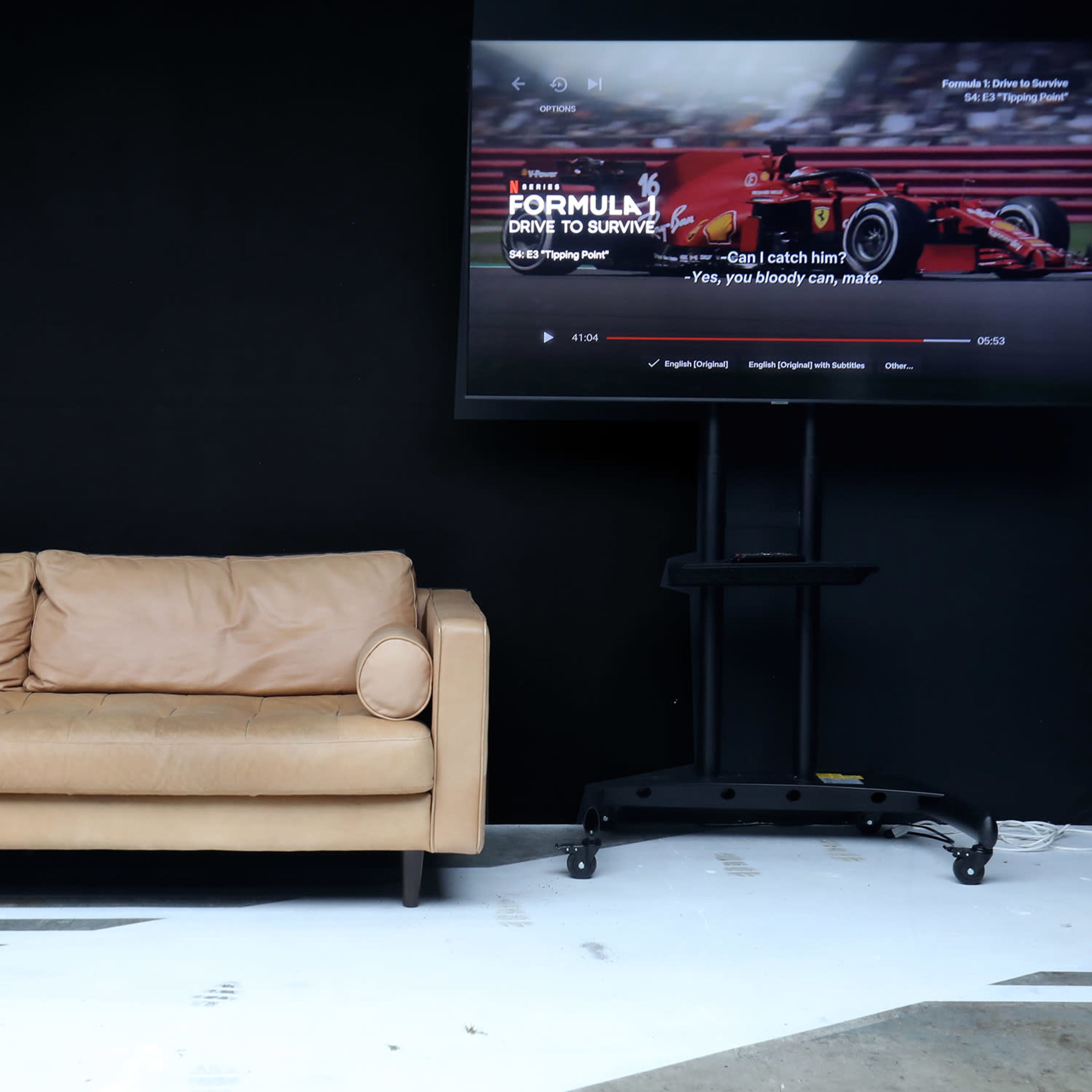 A tan leather couch is positioned in front of a large TV screen displaying a Formula 1 racing program in a minimalist black and white room.