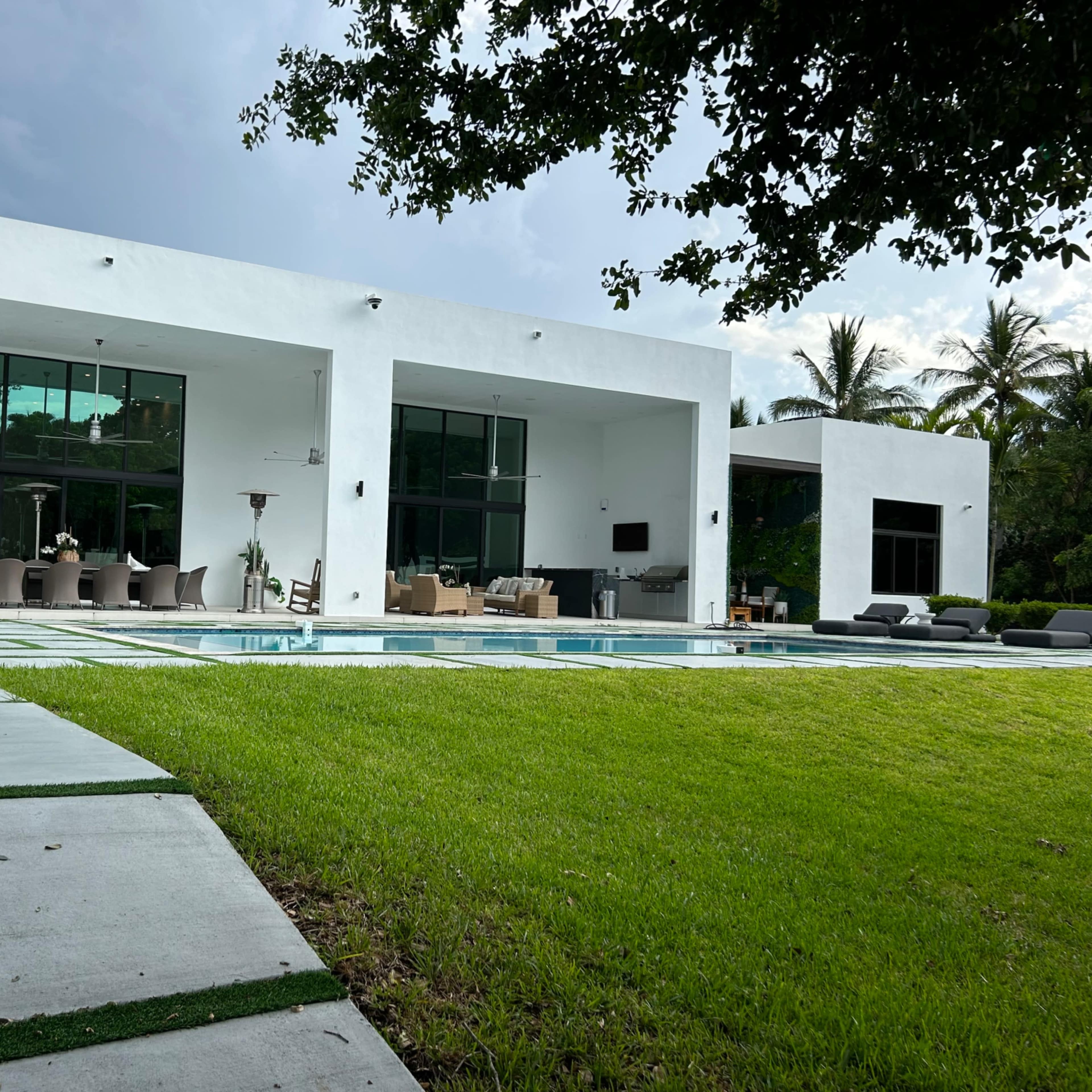 A modern white house with large windows and a swimming pool surrounded by a grassy area and palm trees.