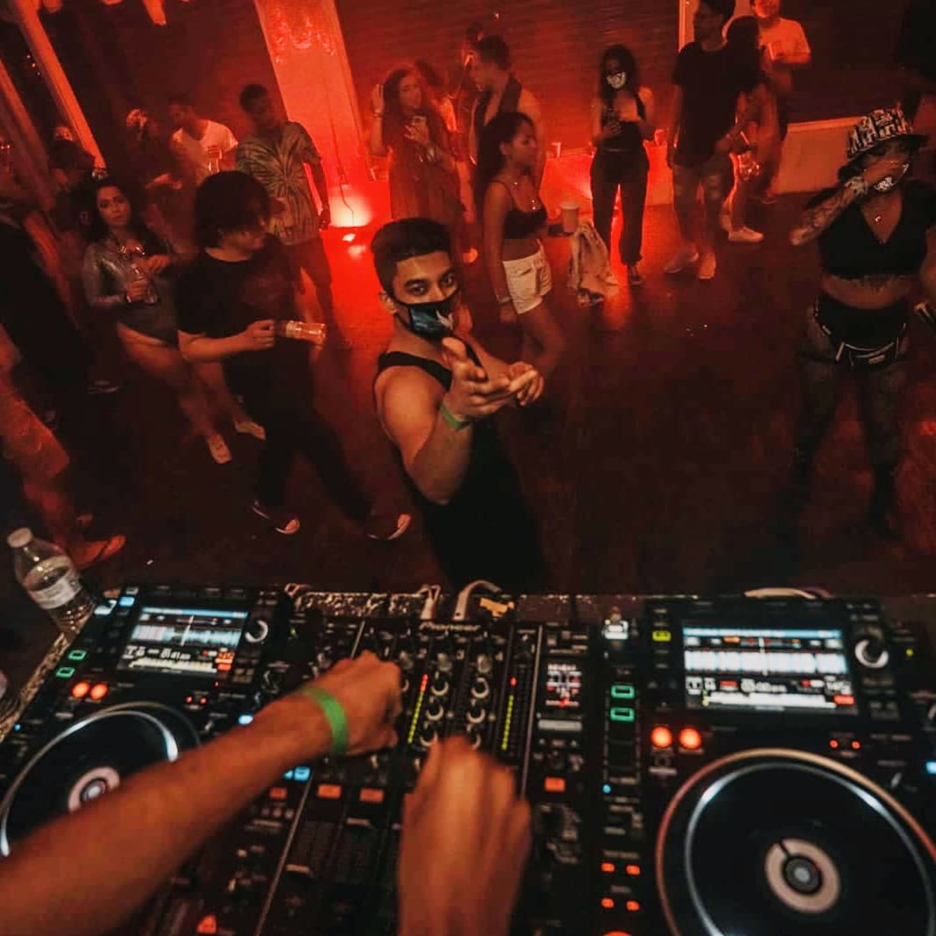 A DJ, with hands on the mixing equipment, performs in a crowded club illuminated by red lights, while partygoers dance in the background.