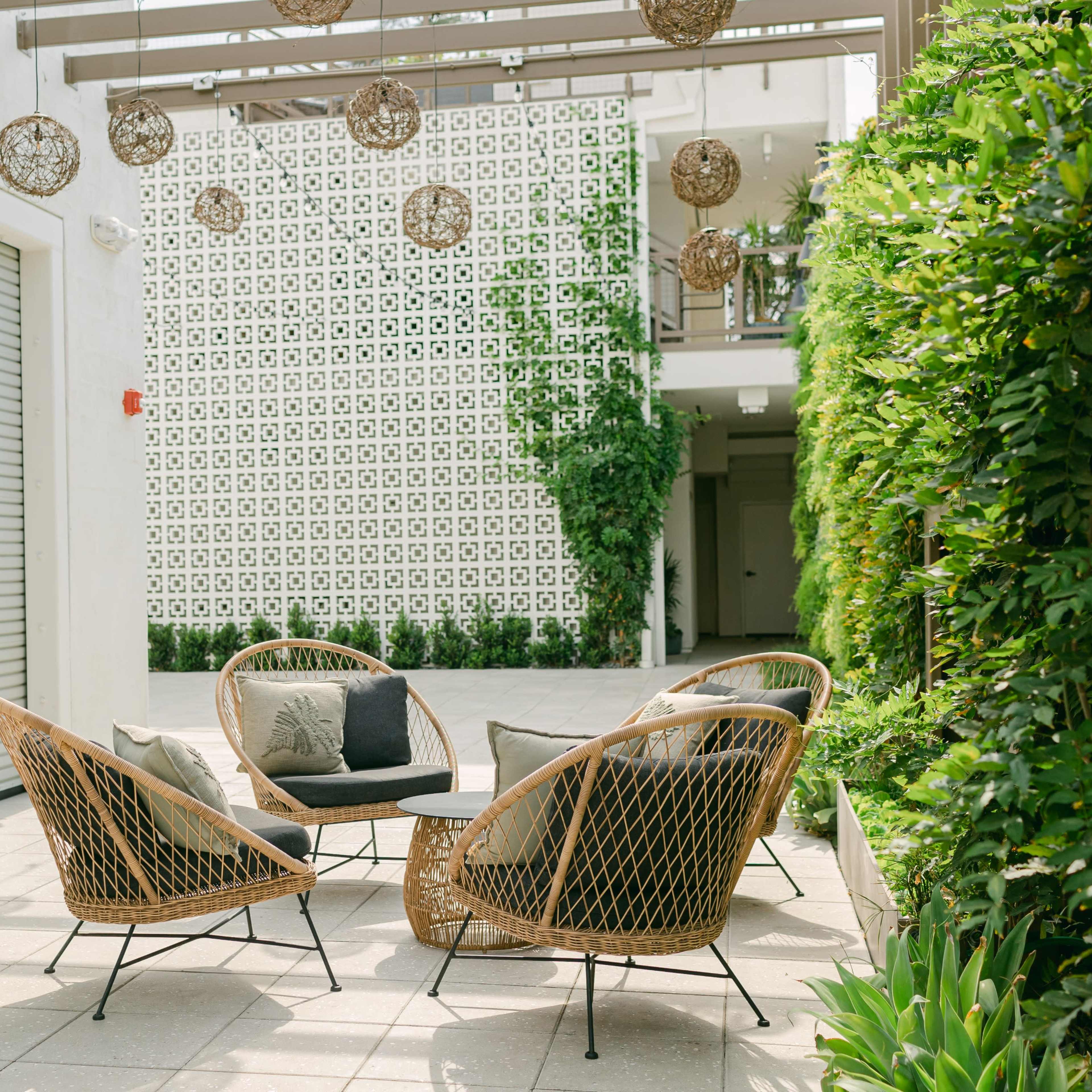A modern outdoor seating area features four woven chairs surrounding a small table, with greenery and a patterned wall in the background.