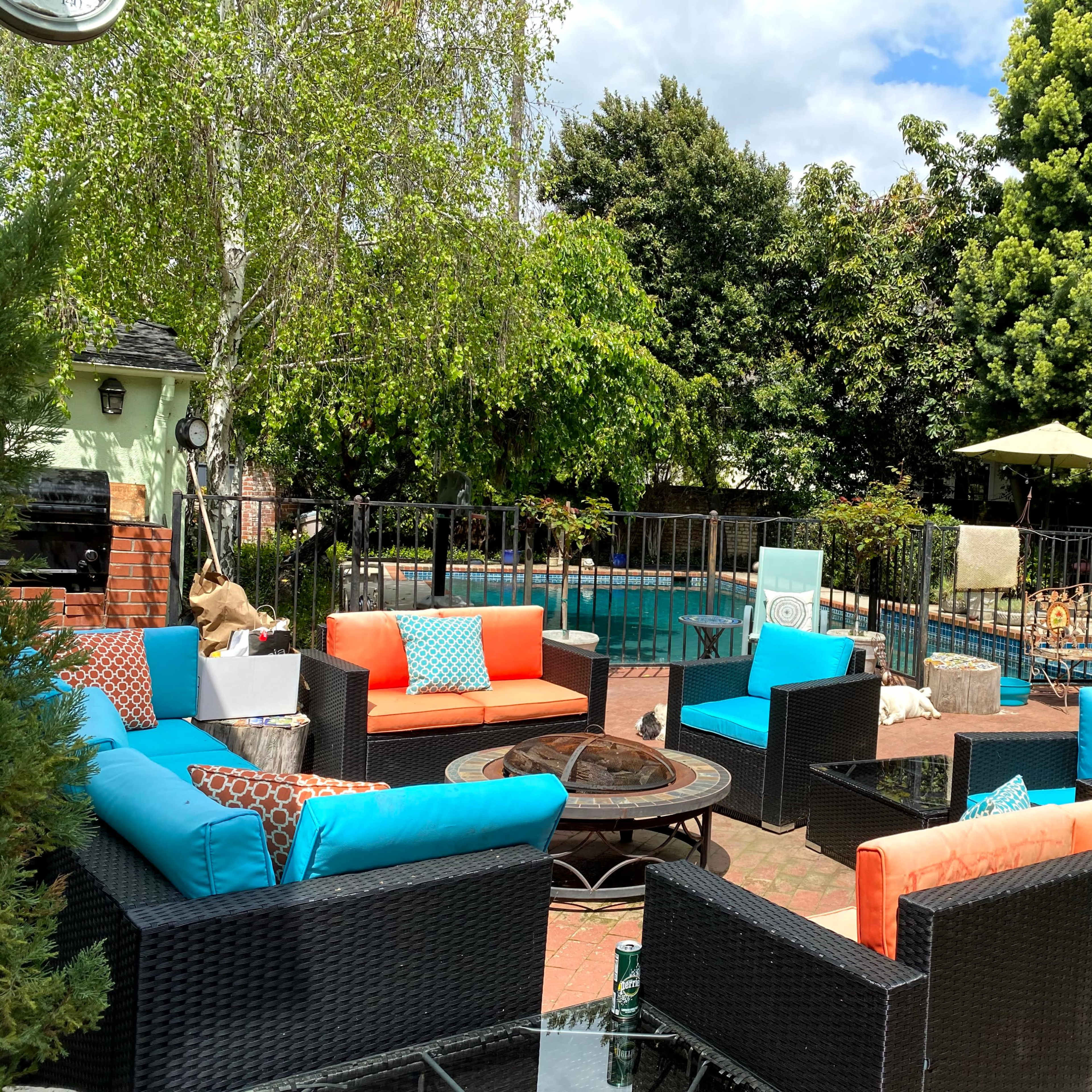 A landscaped backyard with a swimming pool surrounded by seating areas featuring bright blue and orange cushions, along with a fire pit and lush green trees.