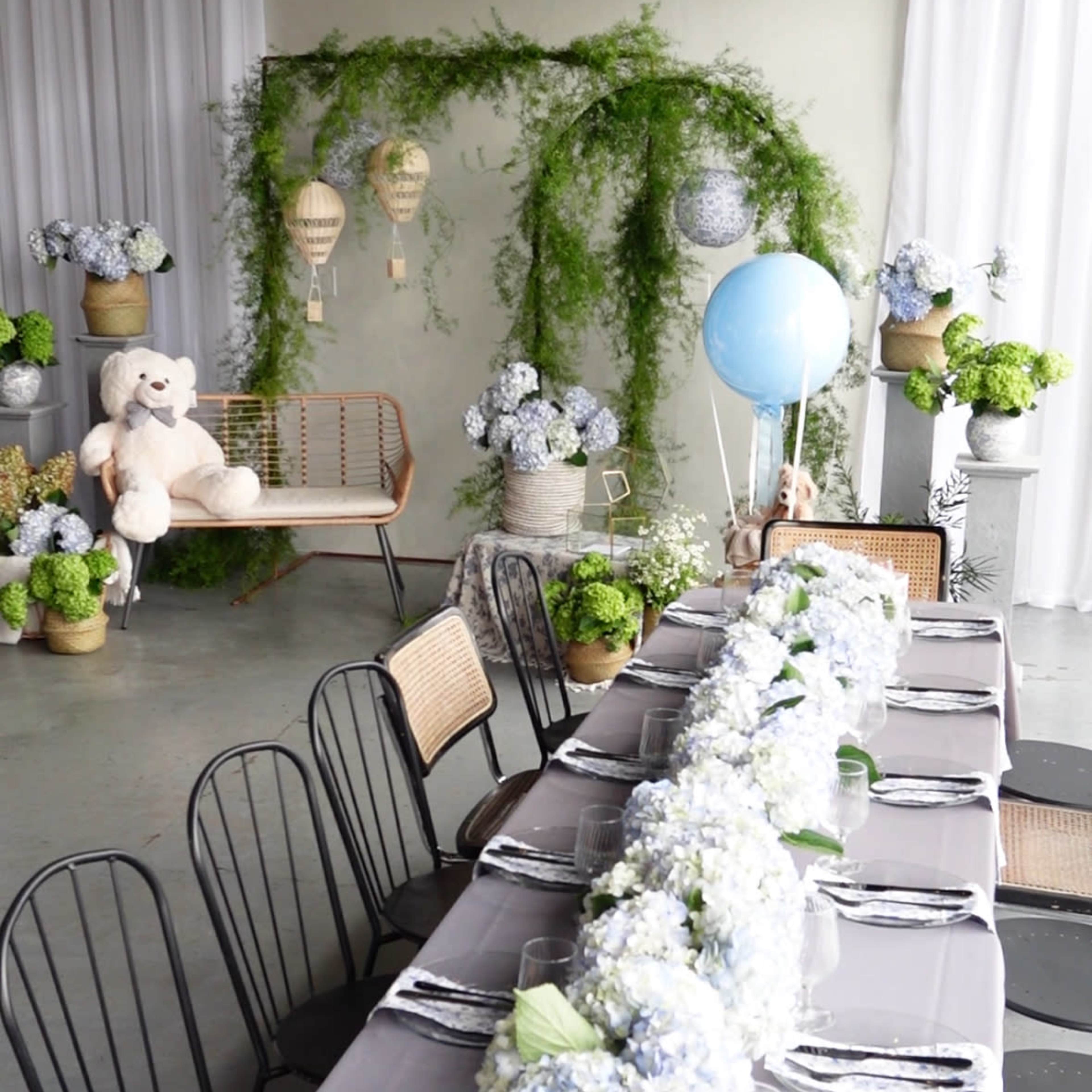 A long dining table adorned with a floral centerpiece is set in a room decorated with greenery, balloons, and a teddy bear against a backdrop of white curtains.