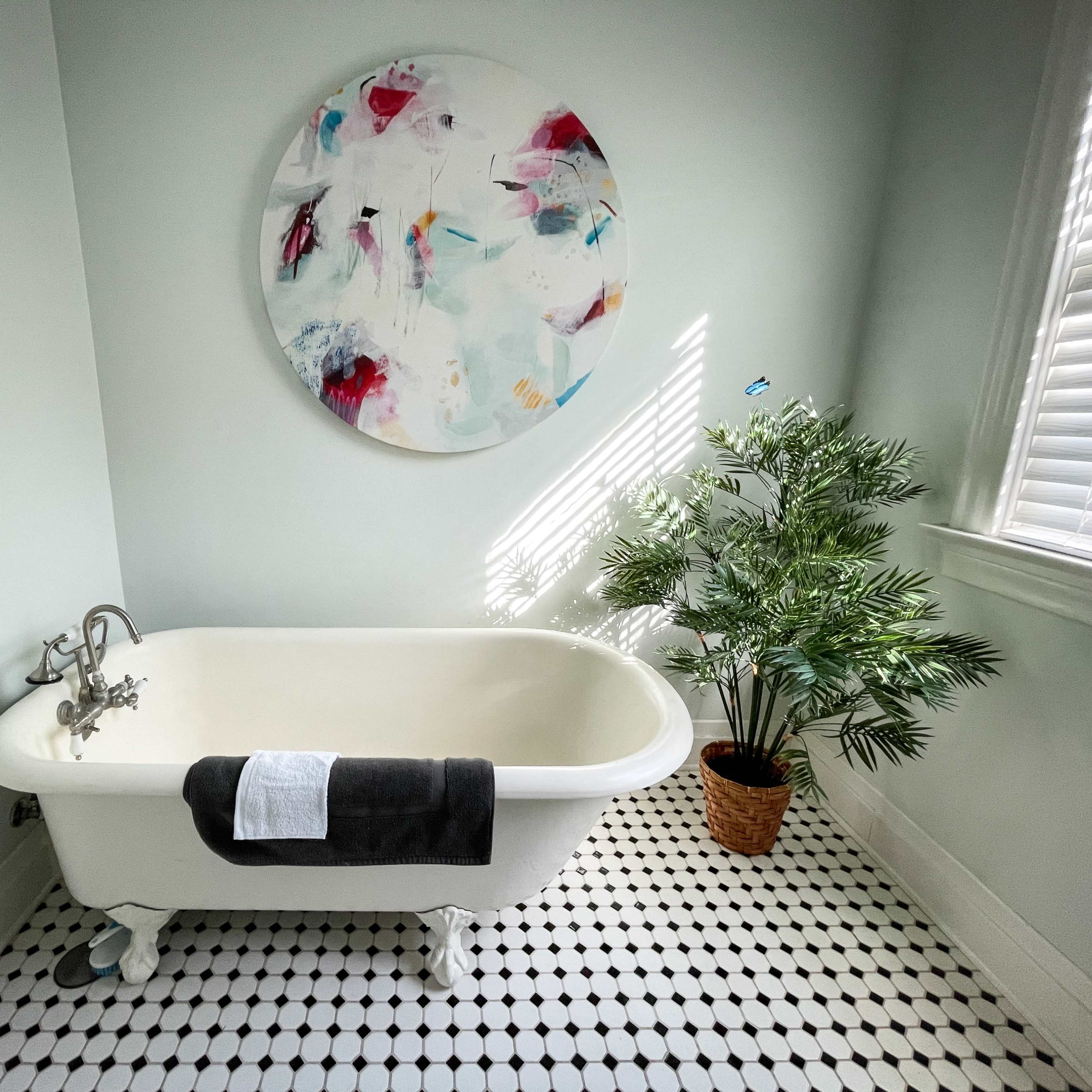 A bathroom featuring a freestanding white bathtub, a round colorful artwork on the wall, and a potted plant beside the tub on a black-and-white tiled floor.