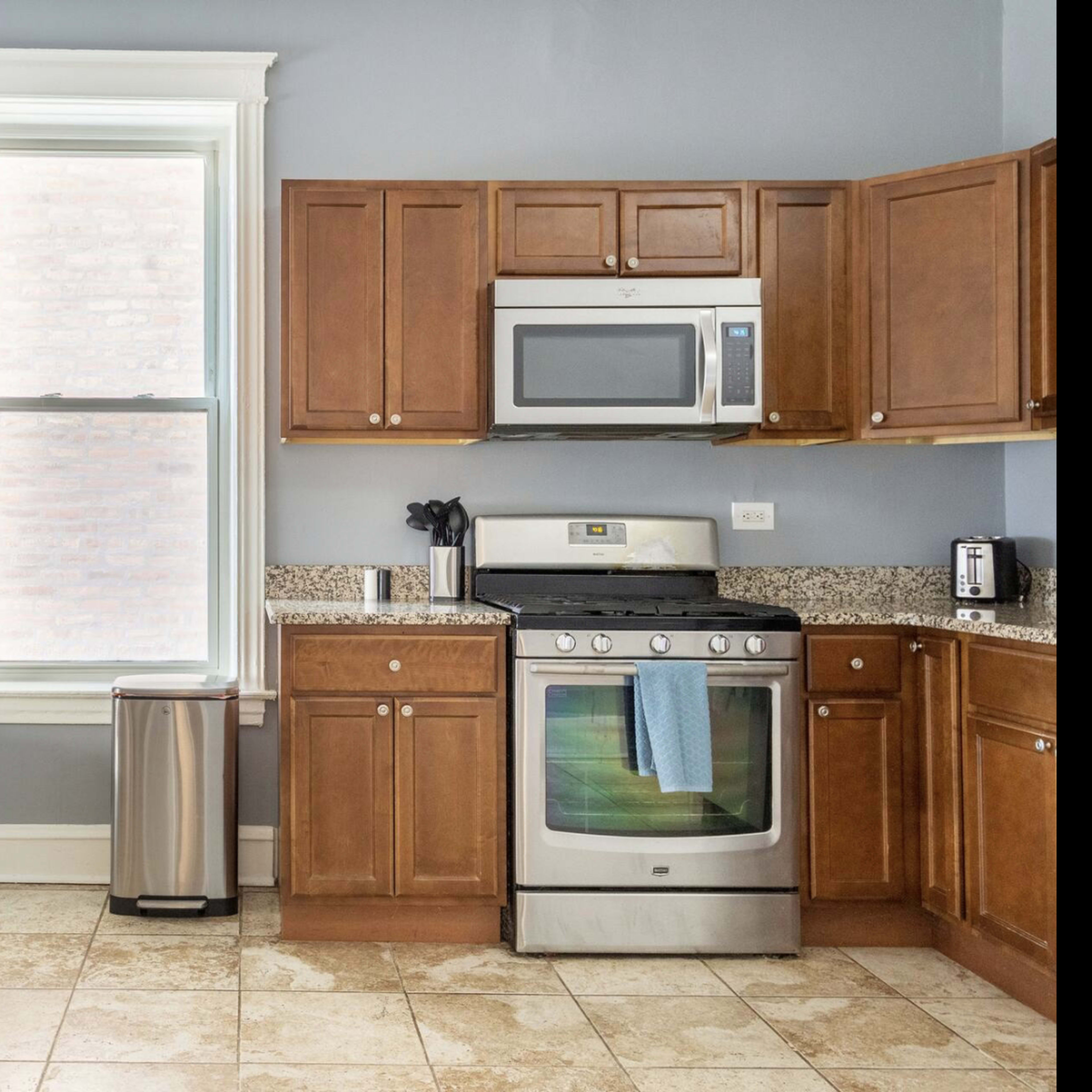 A kitchen features wooden cabinets, a stainless steel stove, a microwave, and a small wooden table with two chairs beside a window.