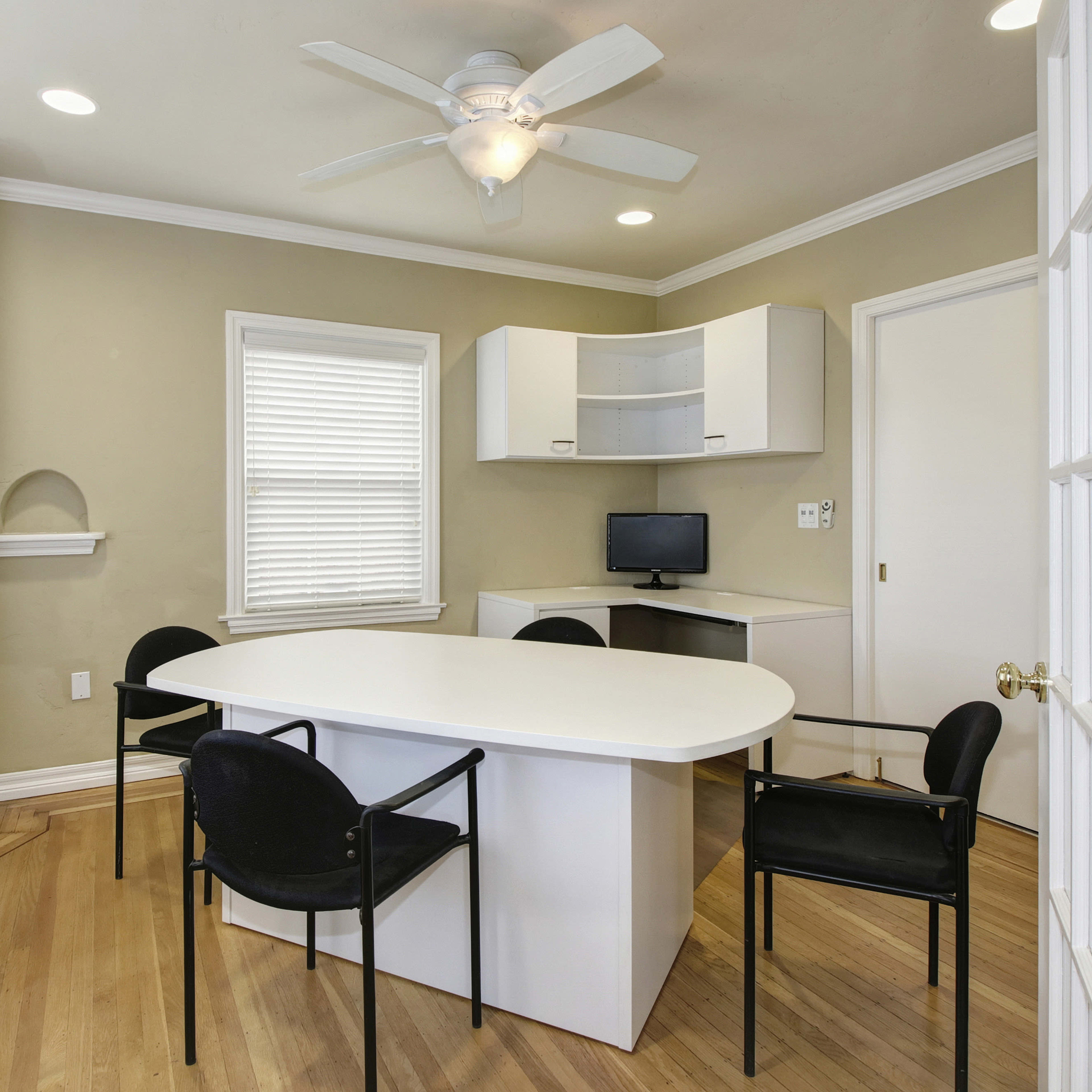 A small workspace with a round table, four black chairs, a wall-mounted desk with a computer, and natural light from multiple windows.