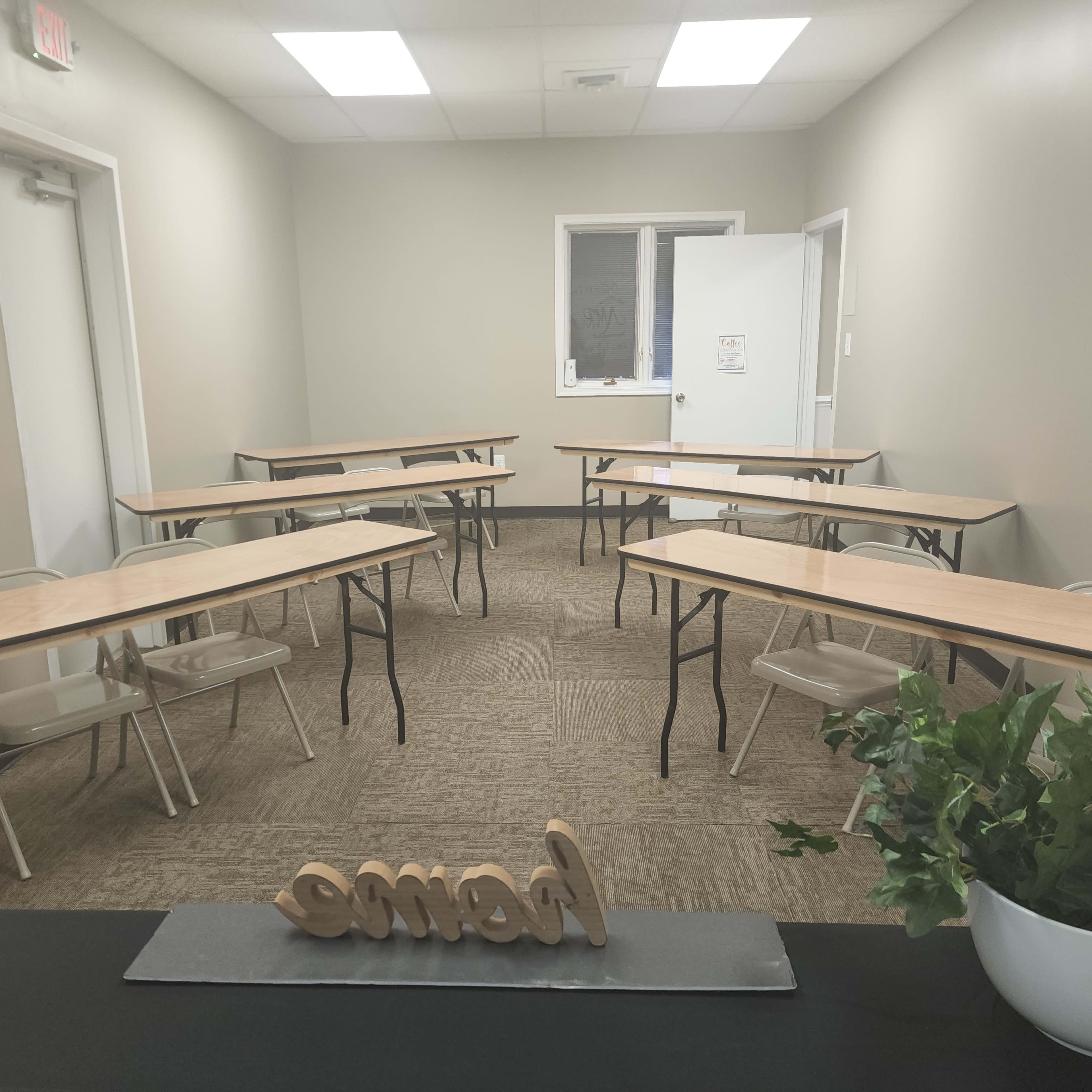 A small, vacant classroom with several tables arranged in a U-shape and a plant on a desk at the front.