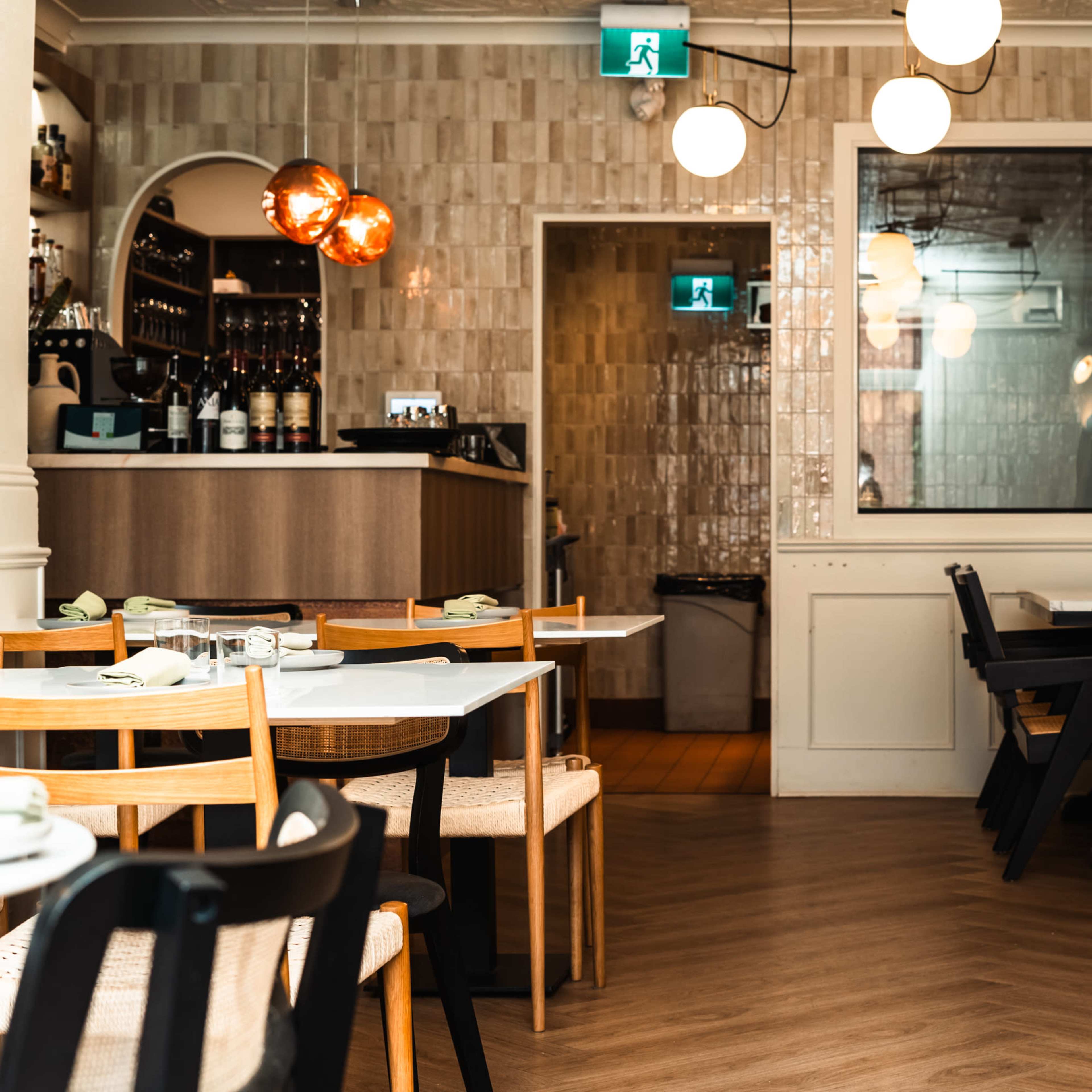 A contemporary restaurant interior with wooden tables and chairs, a bar area, and decorative lighting features.