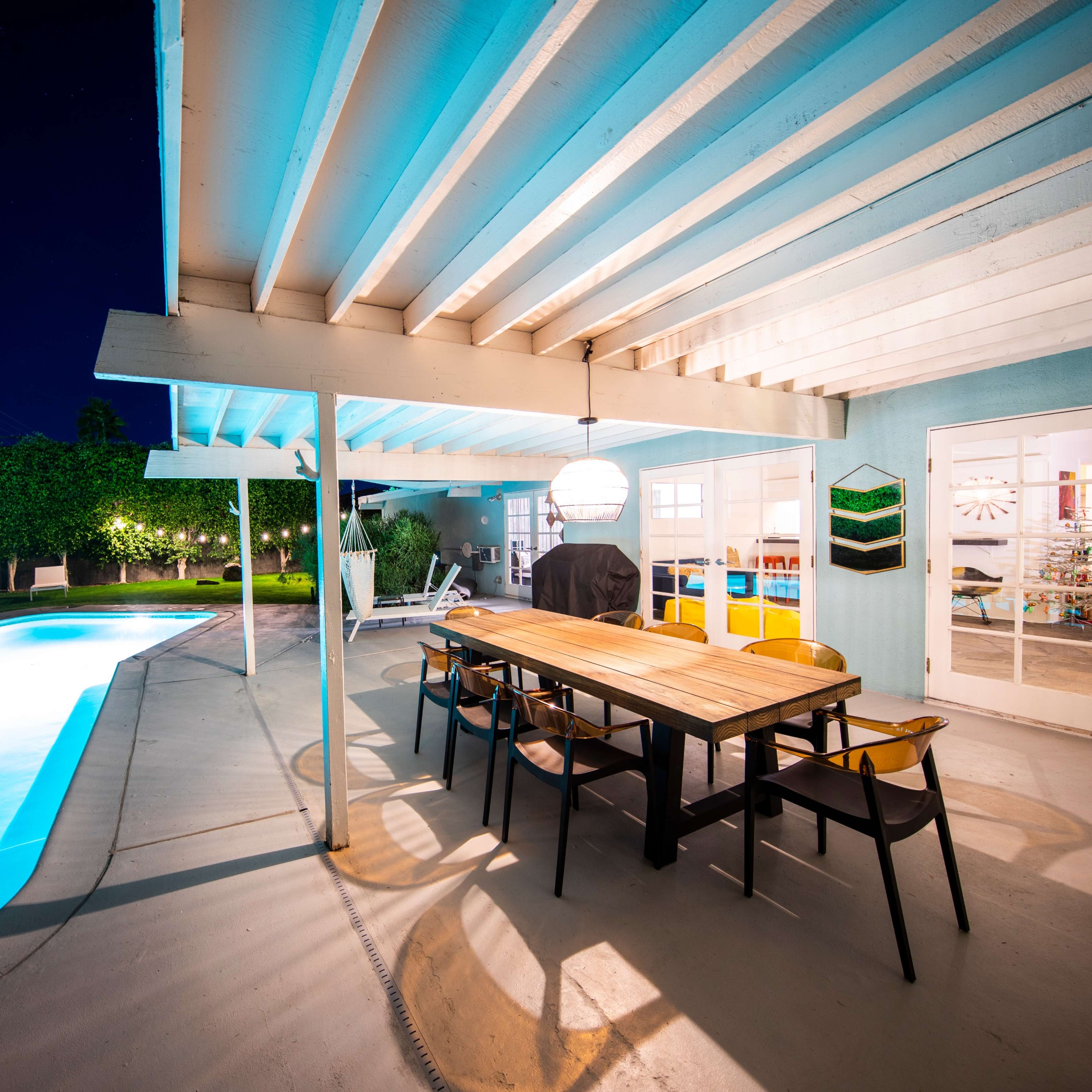 A poolside patio features a long wooden dining table surrounded by chairs, illuminated by a hanging light, with a swimming pool and greenery in the background.