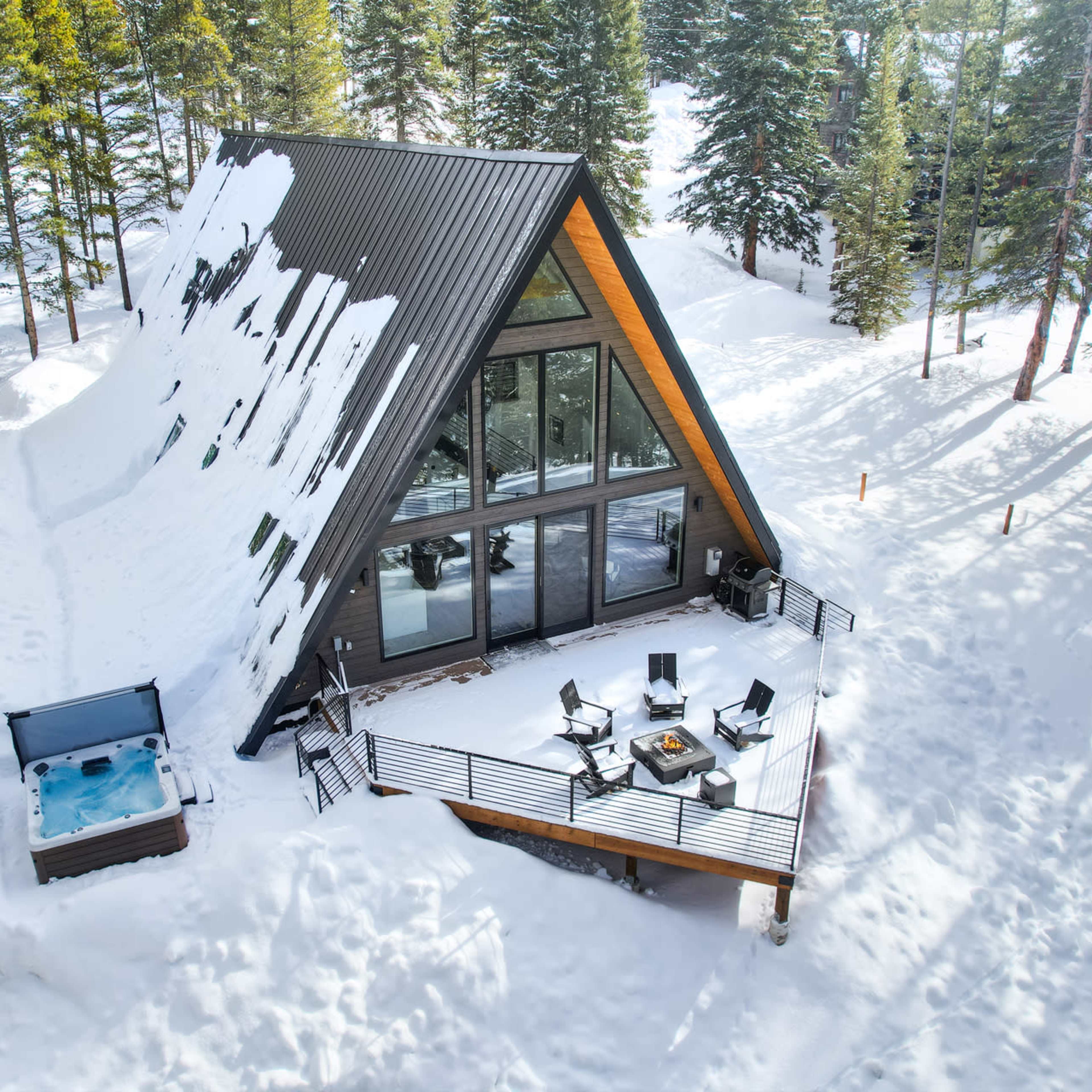 A snow-covered A-frame cabin with large windows and a hot tub is surrounded by tall evergreen trees.
