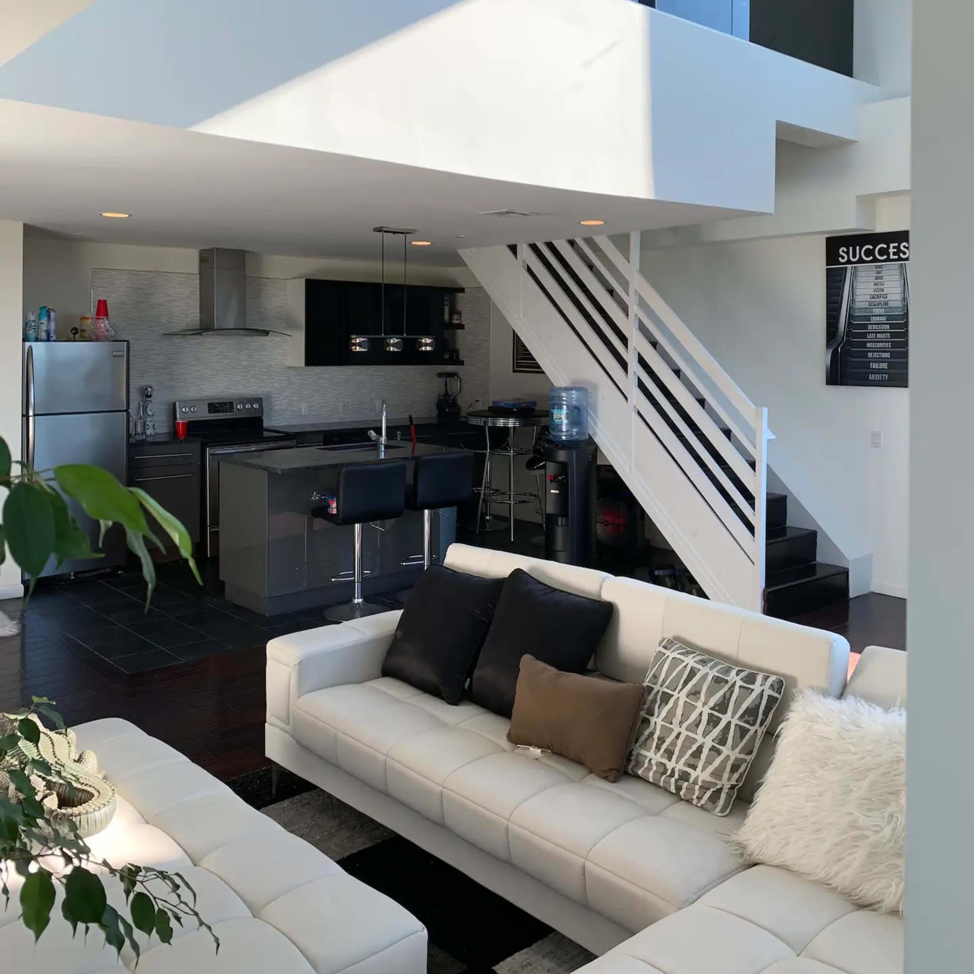 A modern open-concept living space with a white sofa, a kitchen area, and a staircase leading to a lofted area.