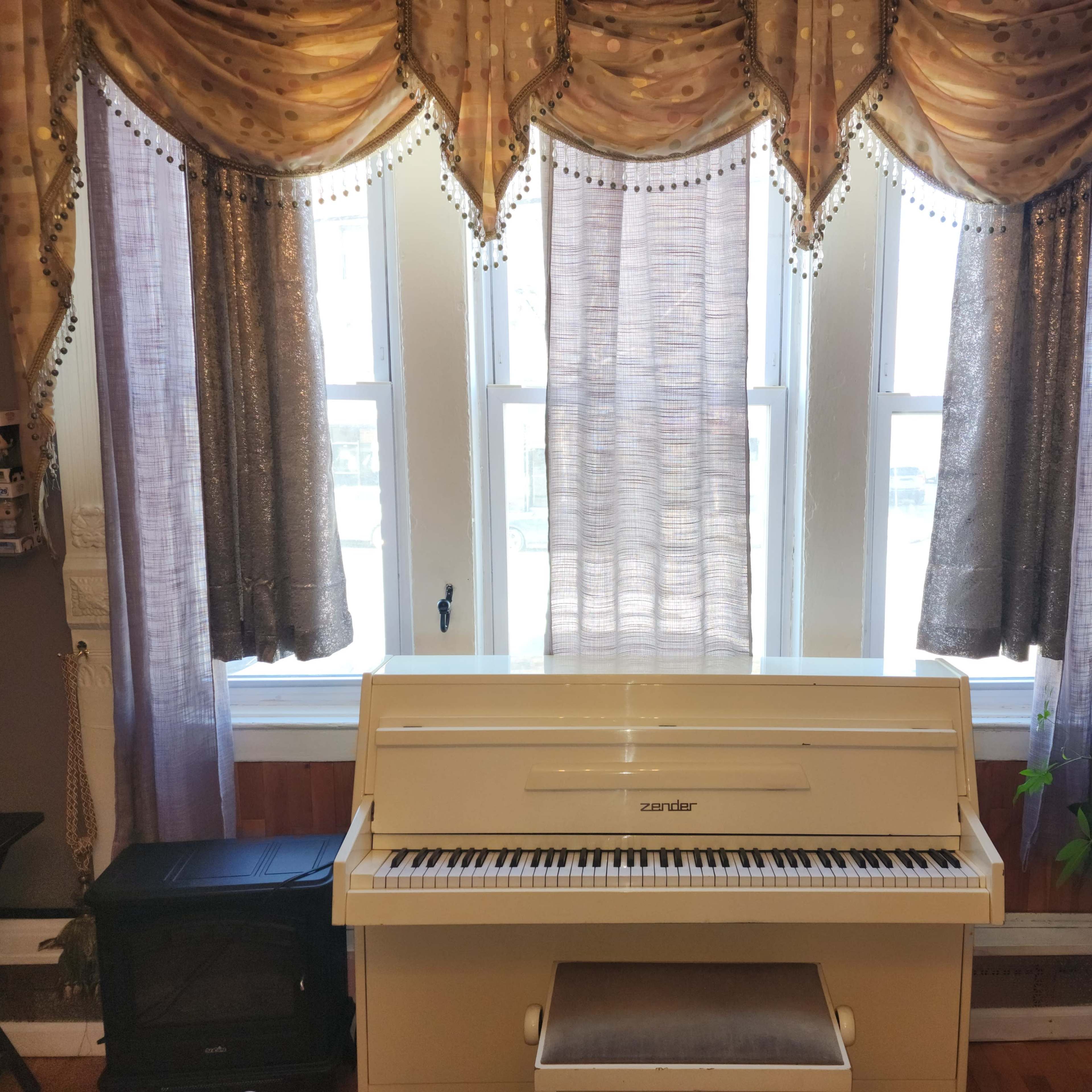 A white piano sits below two large windows adorned with patterned drapes.