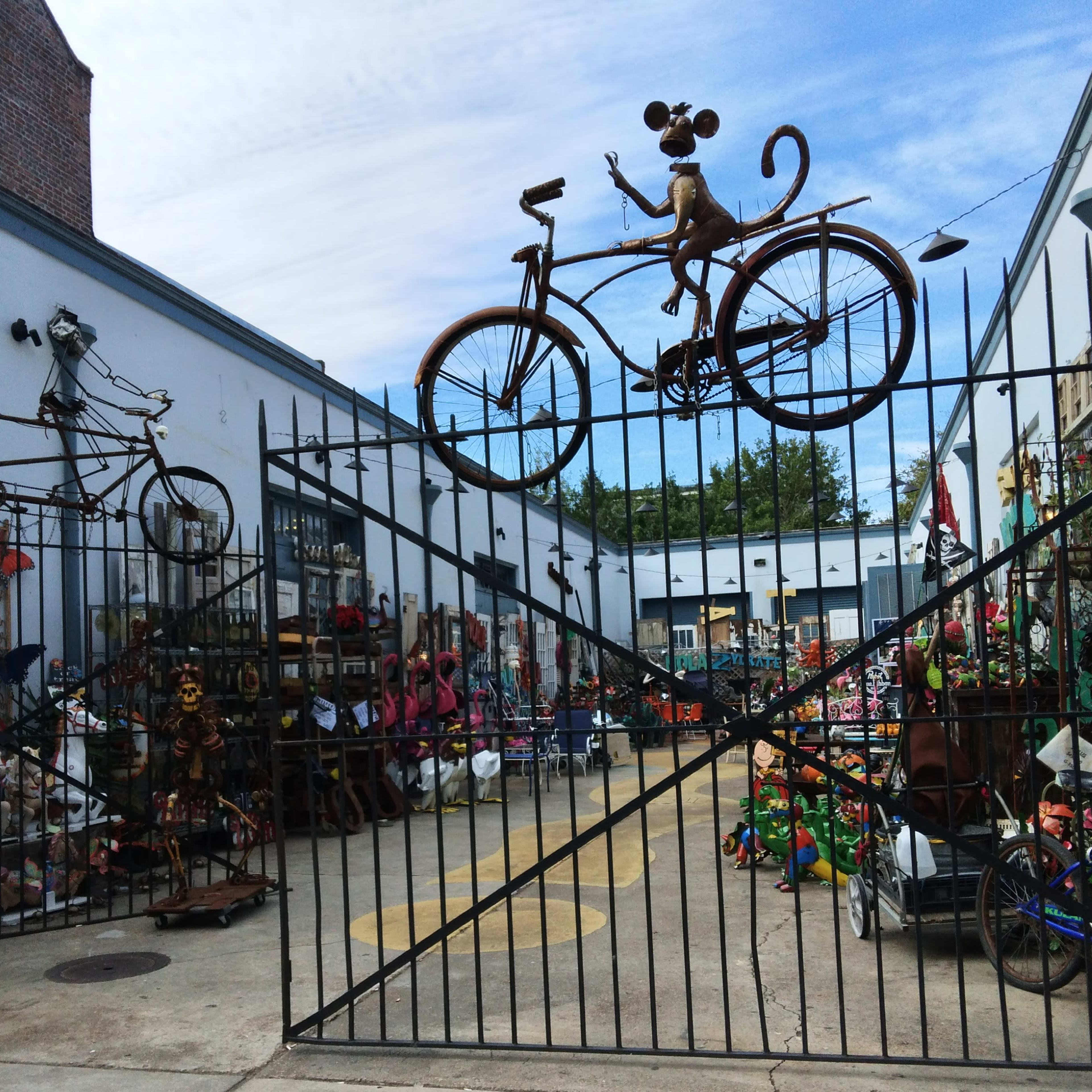 A decorative metal gate with a bicycle and a monkey sculpture on top, leading into a vibrant outdoor space filled with various sculptures and items for sale.
