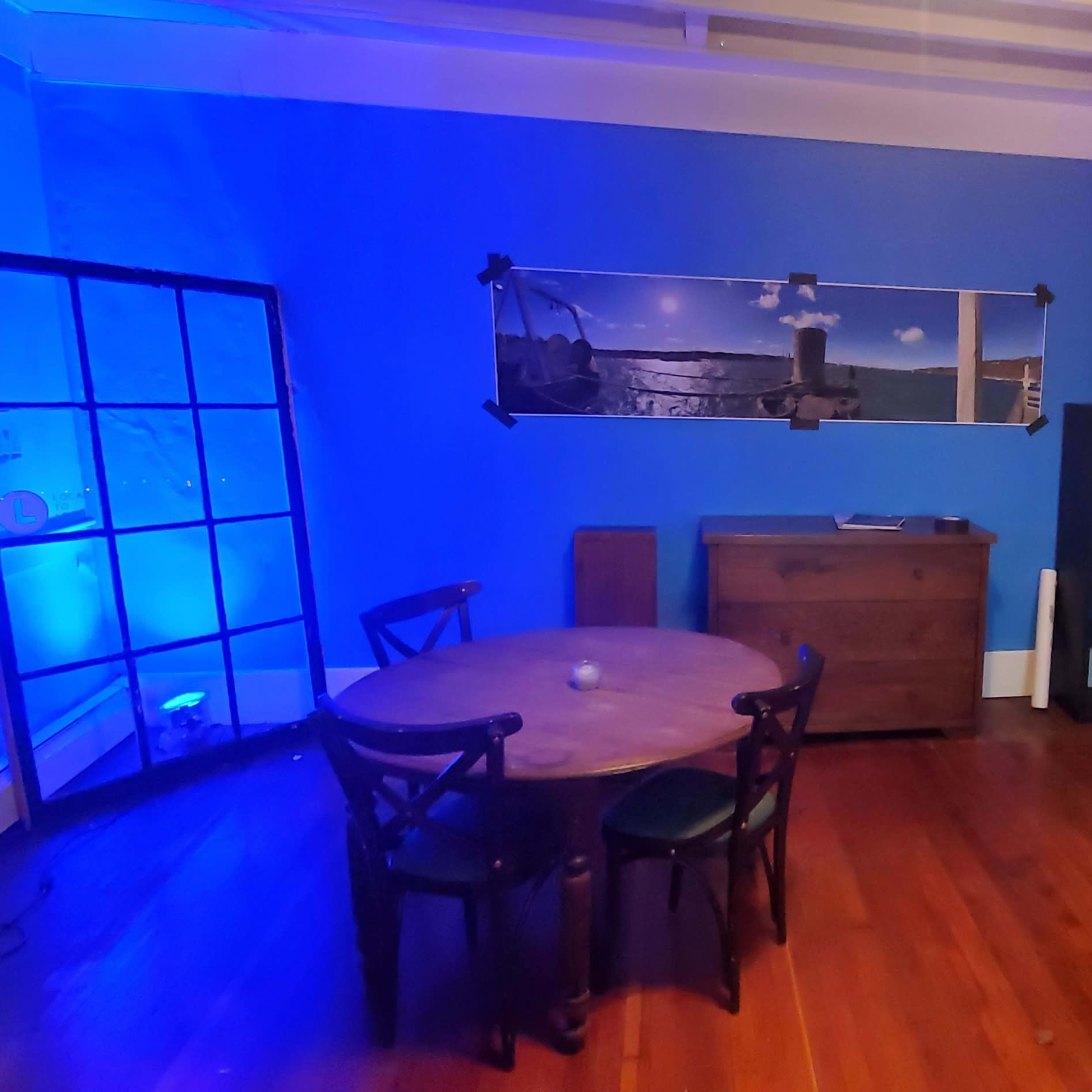 A room featuring a circular wooden table with four chairs, a blue wall with a large photograph, a sideboard, and a bar with high stools.