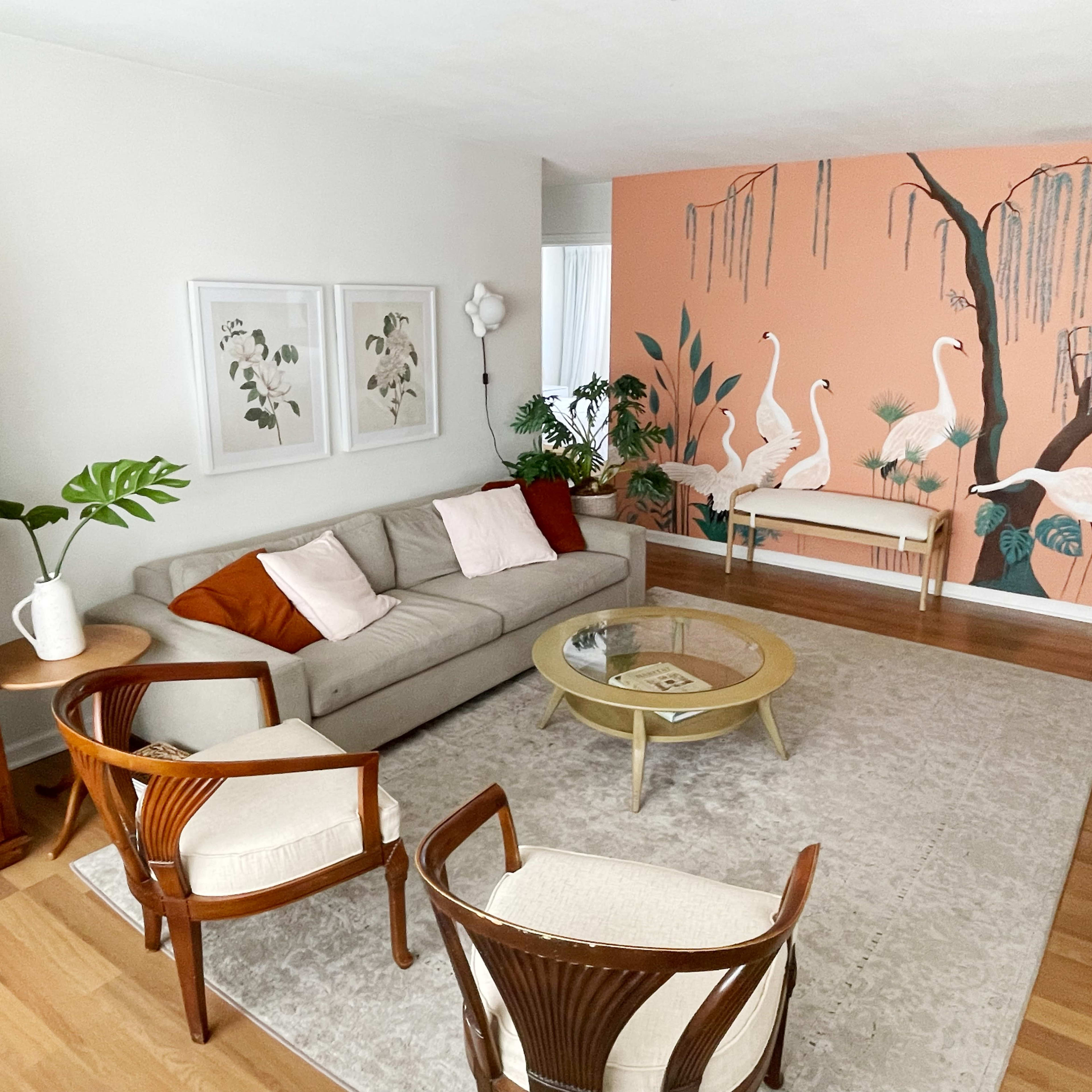 The living room features a light gray sofa with pillows, two wooden chairs, a round coffee table, and a mural of flamingos on a coral wall.
