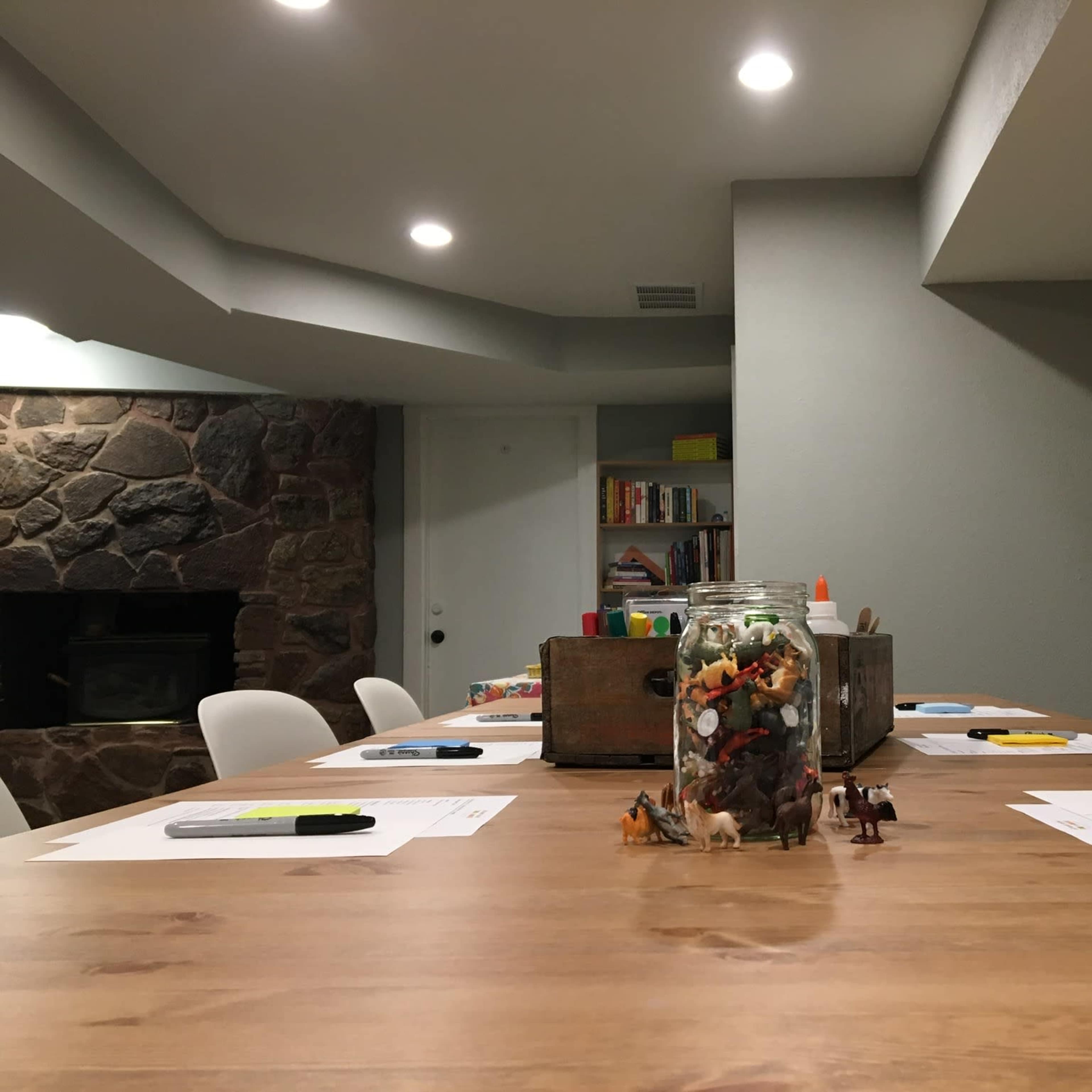 A long wooden table is set up in a well-lit meeting room, with papers and sticky notes placed at each seat and a jar filled with small toys at the center.