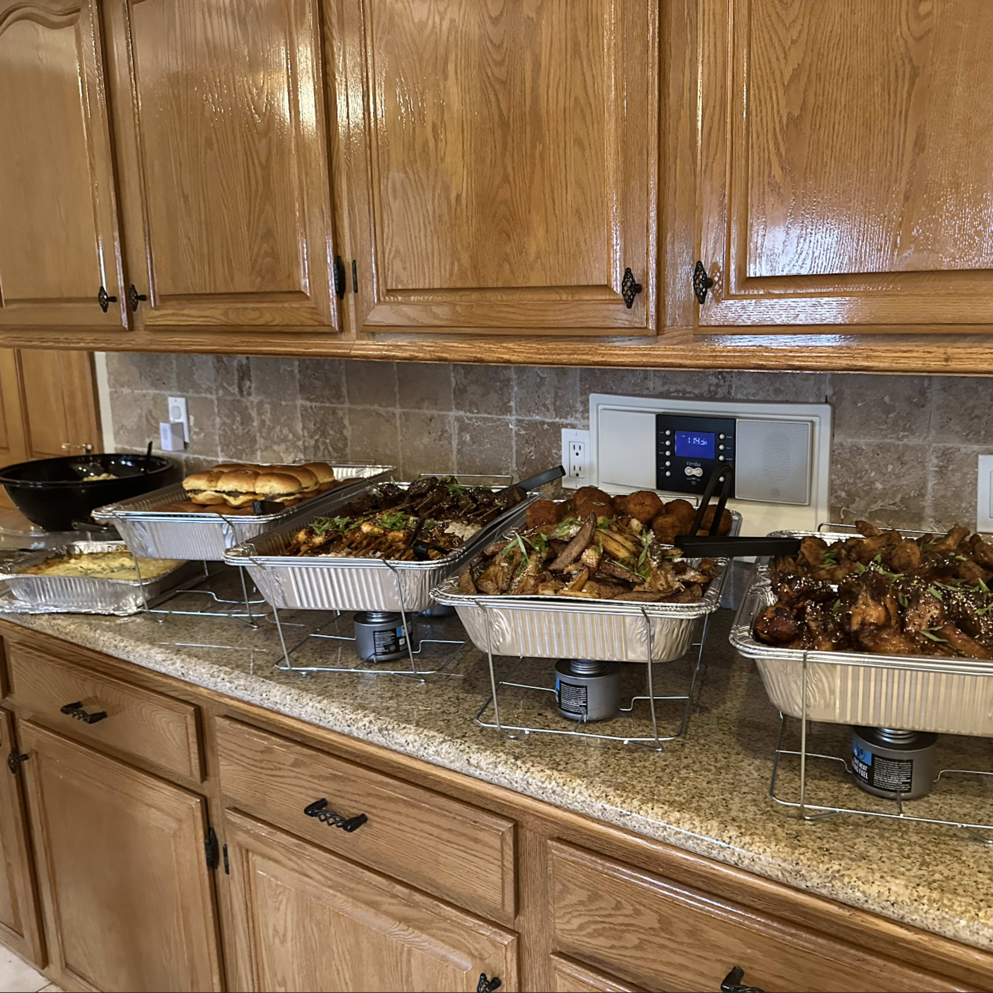 A kitchen counter with several large trays of assorted food, including meats, vegetables, and bread, arranged neatly in front of wooden cabinets.